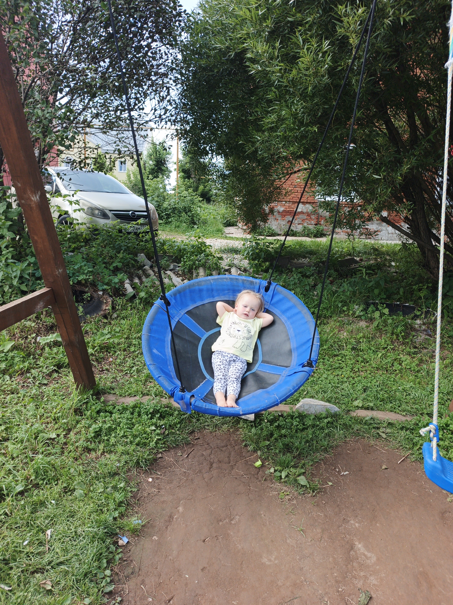 A young child with blonde hair, wearing a light yellow t-shirt with a graphic and patterned leggings, is lying relaxed on a blue tire swing in an outdoor setting. The swing is suspended by black ropes from a wooden A-frame structure. The ground beneath the swing is a mixture of dirt and green grass. In the background, there is dense green foliage, a brick building, and a light-colored car parked amongst the greenery. The overall scene suggests a residential backyard in Medvedyovka, Russia, likely during the daytime.