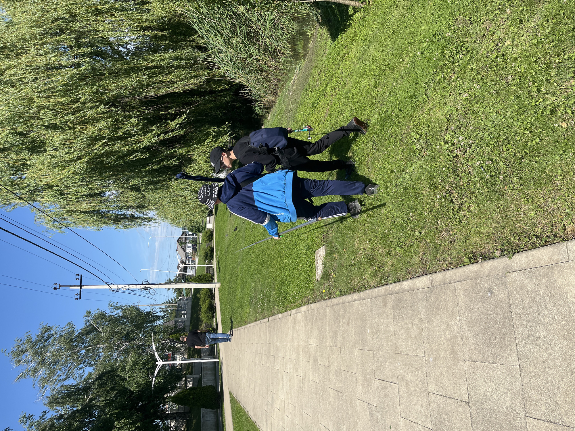Two individuals are engaged in fishing along the bank of a waterway in Goryachiy Klyuch, Russia. One person is wearing a black jacket, dark pants, a black baseball cap, and carrying a backpack. The other person is wearing a blue jacket and dark pants with white stripes. Both figures are positioned close to the edge of the bank. A fishing rod is visible in their hands. The bank is green with grass. In the background, there is a sidewalk, and a man in a black shirt and blue jeans is walking along the sidewalk. Trees, telephone poles, and buildings are also visible in the distance. The sky is blue.