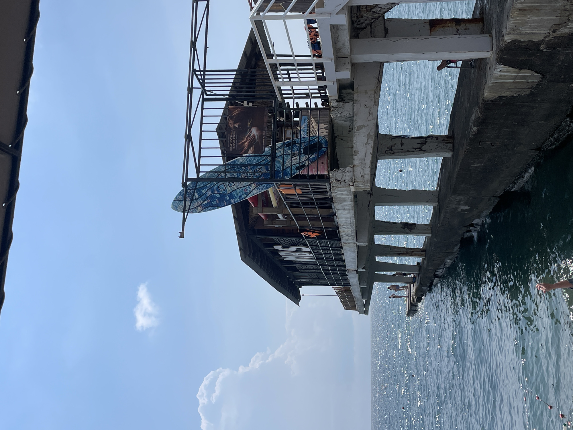The image depicts a pier extending into the Black Sea in Sochi, Russia. A weathered, open-air structure, likely a cafe or bar, is situated on the pier, supported by concrete pillars. The building has a thatched roof and wooden walls with the letters "MBS" prominently displayed. A surfboard is propped against a railing in front of the building. A poster with Russian text is also visible on the structure. Several people are present on the pier and in the water. The sea is choppy with sunlight reflecting off the surface. The sky is blue with scattered clouds, one large cumulus cloud dominating the left side of the frame. The overall scene suggests a recreational area by the sea on a sunny day.