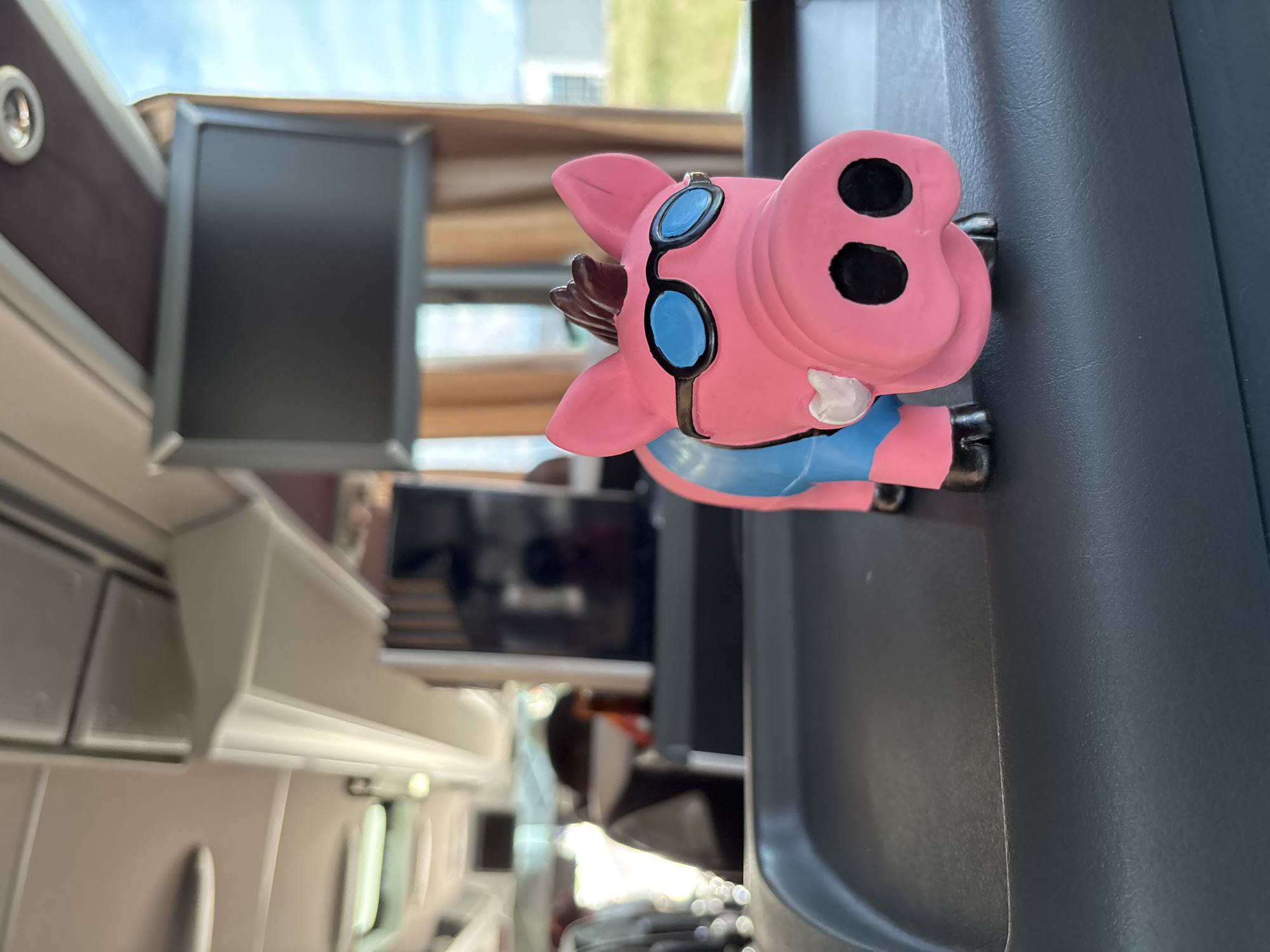 The image shows a pink pig figurine wearing blue goggles and a blue shirt, placed on a gray surface inside a train. The train appears to be a high-speed train, based on the interior design, possibly an Italo train. In the background, there are other train seats, overhead compartments, and a small monitor. Through the windows, glimpses of greenery and buildings are visible, suggesting a location in the city of Prato, Italy.