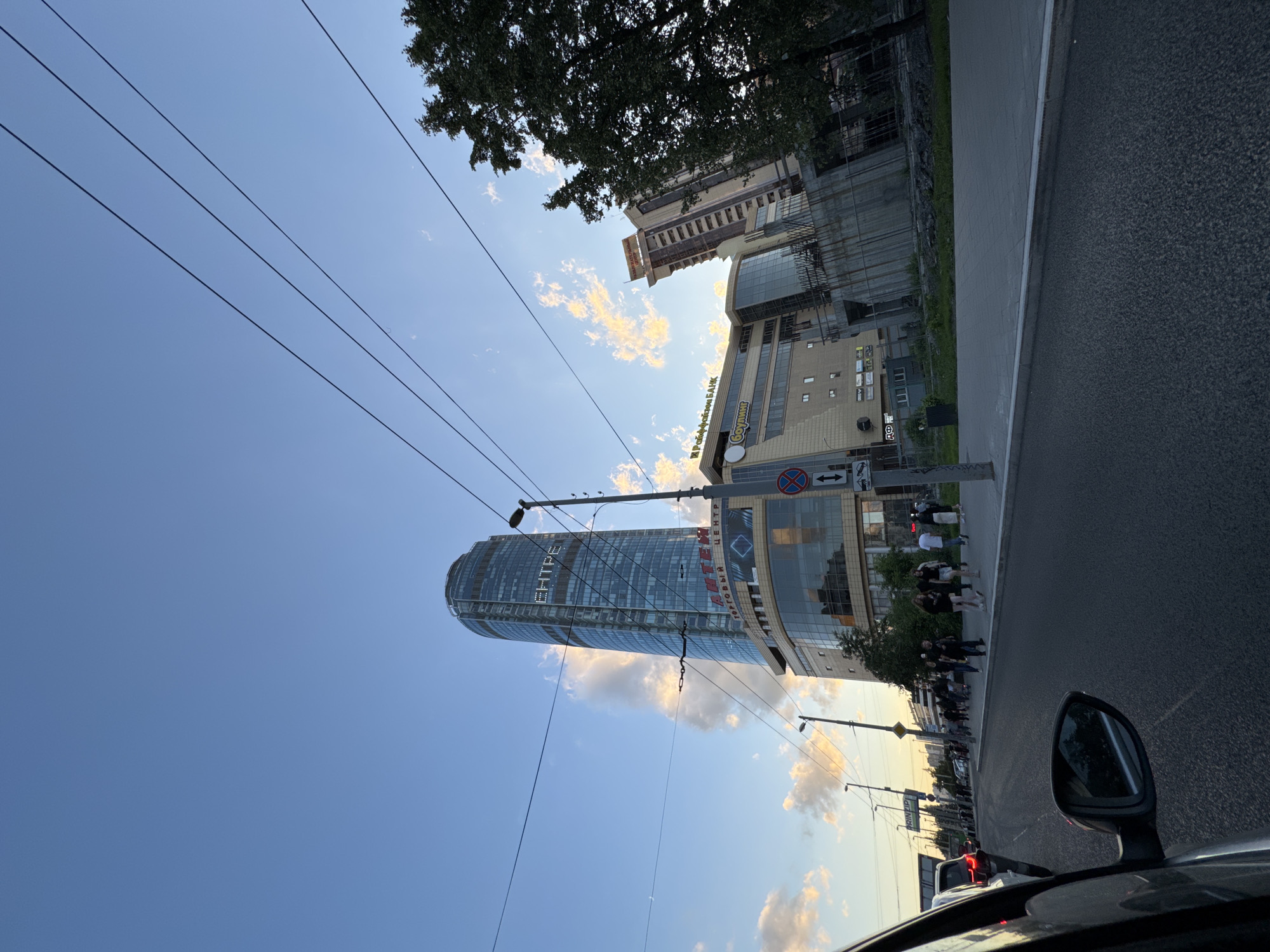 The image shows a street scene in Yekaterinburg, Russia, focusing on a prominent glass skyscraper named "Vysotsky" with the Cyrillic text "ЕЦНТРЕ" visible on its facade, which translates to "CENTER". The skyscraper is a modern, tall building with a distinctive rounded top. To the left of the skyscraper is a lower, multi-story building with extensive glass windows and signage, including the text "ТОРГОВЫЙ ЦЕНТР" ("SHOPPING CENTER"). Further to the right, another tall building with a more beige facade rises, featuring visible windows and balconies. The foreground shows a street with traffic, with the side mirror and part of a car visible on the left. A sidewalk runs alongside the street, populated by several pedestrians walking in the same direction. Streetlights and overhead power lines are present, indicating urban infrastructure. The sky is a clear blue with scattered, soft clouds tinged with the warm light of what appears to be late afternoon or early evening. The overall impression is of a busy urban environment with modern architecture under a pleasant sky.