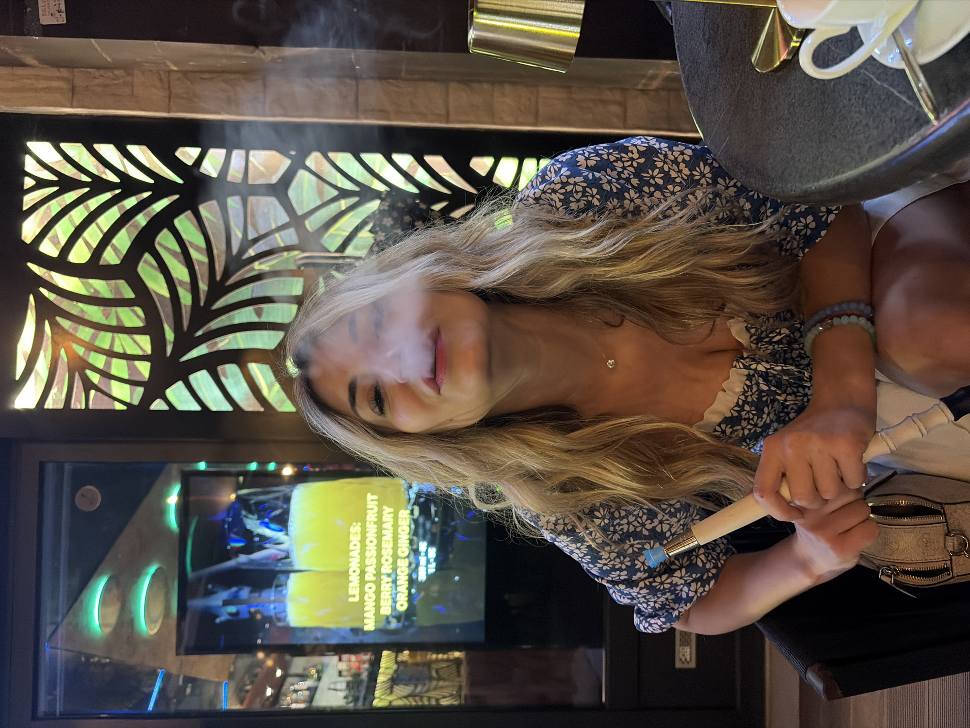 The image depicts a woman with blonde hair smoking, likely a hookah, in a bar or lounge. Smoke emanates from her mouth. She wears a blue and white floral top and has bracelets on her right wrist. In the background, there's a decorative screen with plant-like cutouts and a television displaying a menu of lemonade flavors: mango passionfruit, berry rosemary, and orange ginger. Based on the context (the city of Budva, Montenegro), it's plausible the bar or lounge caters to tourists.