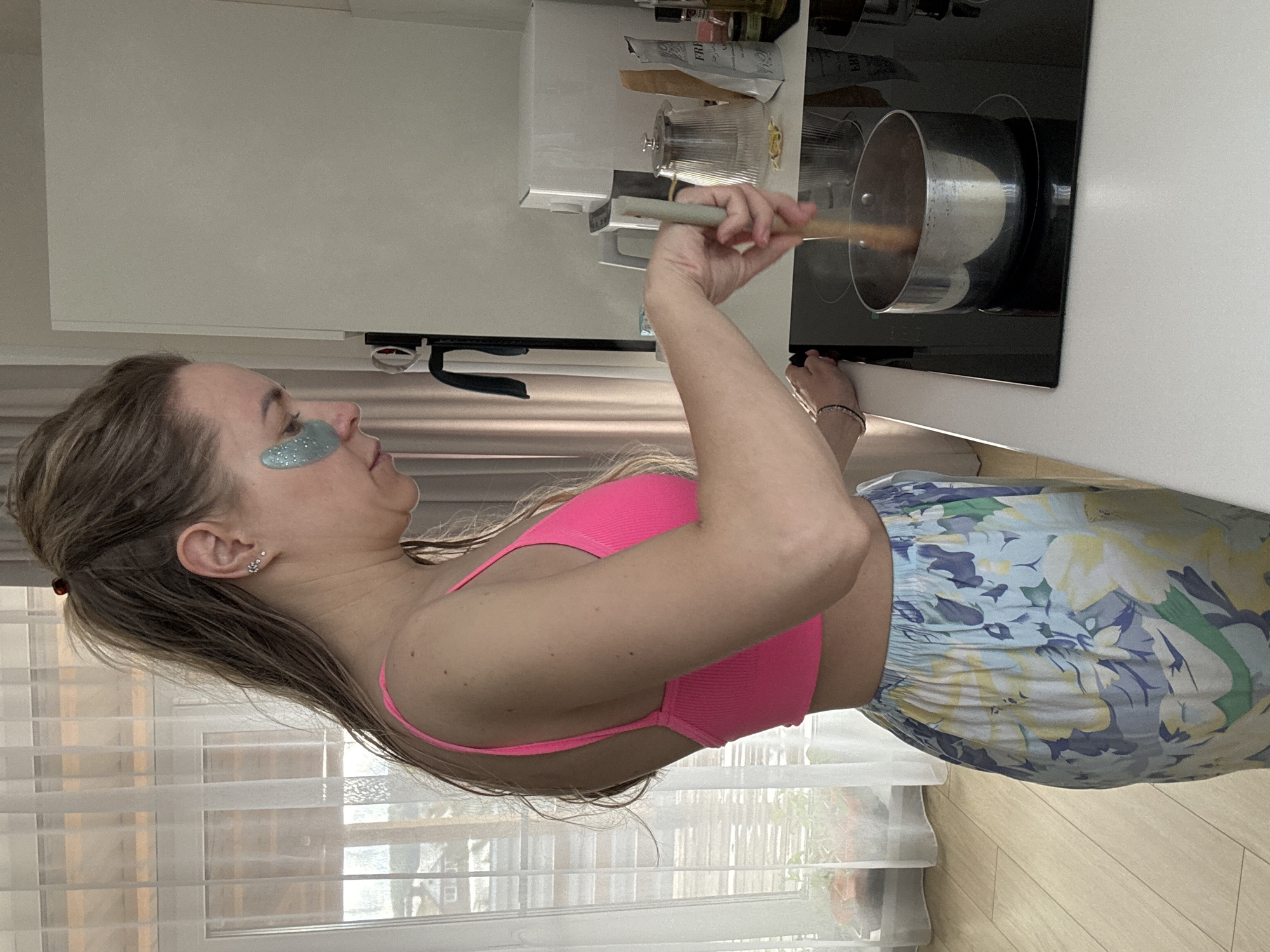 A woman with shoulder-length brown hair pulled back and wearing teal, glittery eye patches stands in a kitchen. She is wearing a bright pink sports bra and blue and yellow floral print pants. She is stirring a pot on a black glass stovetop with a light-colored wooden spoon. The stove is set into a white countertop. White cabinets are above the stove. The scene appears to be inside a residence, evidenced by window blinds and wood flooring. There is no specific evidence to suggest the scene is in Chaikovsky, Russia.