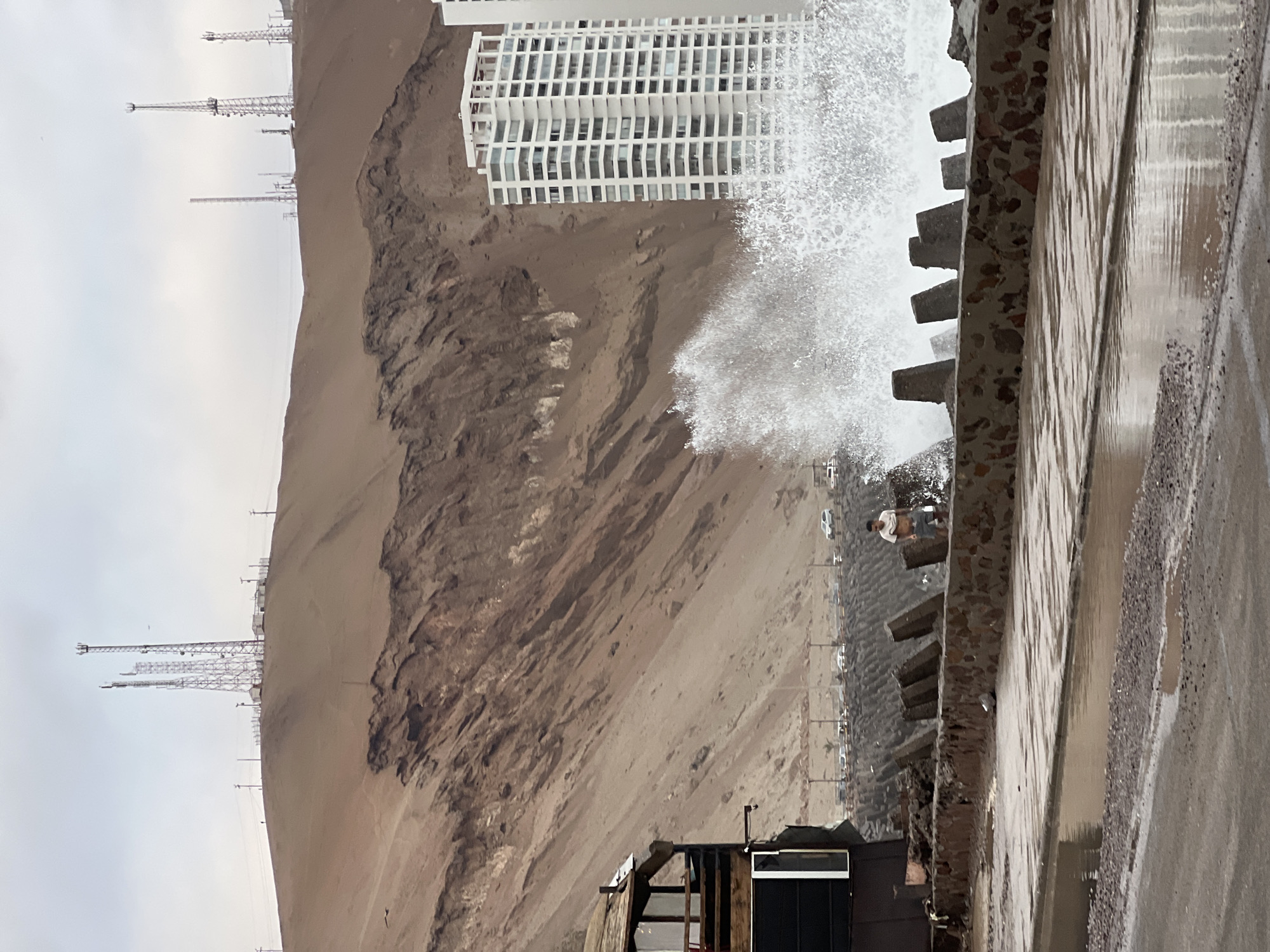 The image depicts a rugged coastline in Arica, Chile, characterized by a steep, sandy hillside topped with communication towers. In the foreground, a concrete seawall, constructed with rough stones and concrete blocks, buffers the shore against the powerful ocean. A large wave is captured mid-break, crashing against the wall with dramatic force, sending spray high into the air. A single individual, a male wearing a light-colored t-shirt and shorts, stands near the seawall, seemingly observing the turbulent sea. To the left, a dark, multi-story building with a weathered wooden facade partially obscures the view of the coastal road, where several vehicles are visible. Further to the right, a tall, white, modern residential building stands prominently against the muted sky, its numerous windows reflecting the overcast conditions. The ground in the immediate foreground is wet and appears to be a pathway or road, with puddles of water reflecting the surrounding environment. The overall scene conveys a sense of raw natural power meeting urban development along the coast of Arica.
