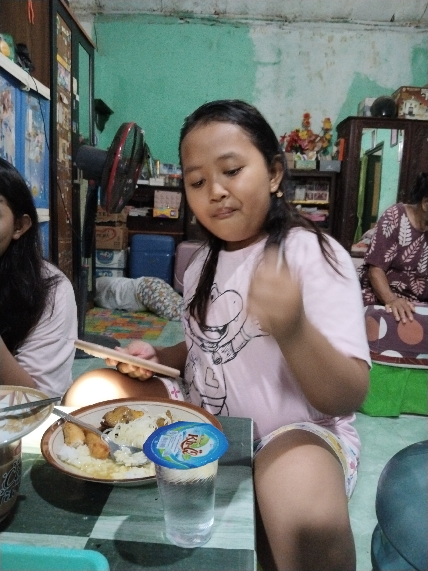 The image depicts an indoor scene, likely a residential interior, in Kecamatan Pati, Indonesia. A young girl, with dark hair and wearing a pink t-shirt with a cartoon graphic and patterned shorts, is seated at a table. She is holding a tablet or similar flat electronic device in her hands. In front of her is a plate of food, consisting of rice, noodles, and what appear to be fried pieces of food. A glass of water with a blue lid from a beverage bottle is also on the table.
To the left of the girl, another child, partially visible, is also seated. In the background, several objects are present, including a black and red electric fan, stacks of boxes, a blue plastic container, and a wooden cabinet with shelves containing various items. A patterned mat or rug is on the floor, and another person is seated further in the background, to the right of the frame, wearing a patterned shirt. The walls are painted a light green color, and the overall environment appears to be a living space.