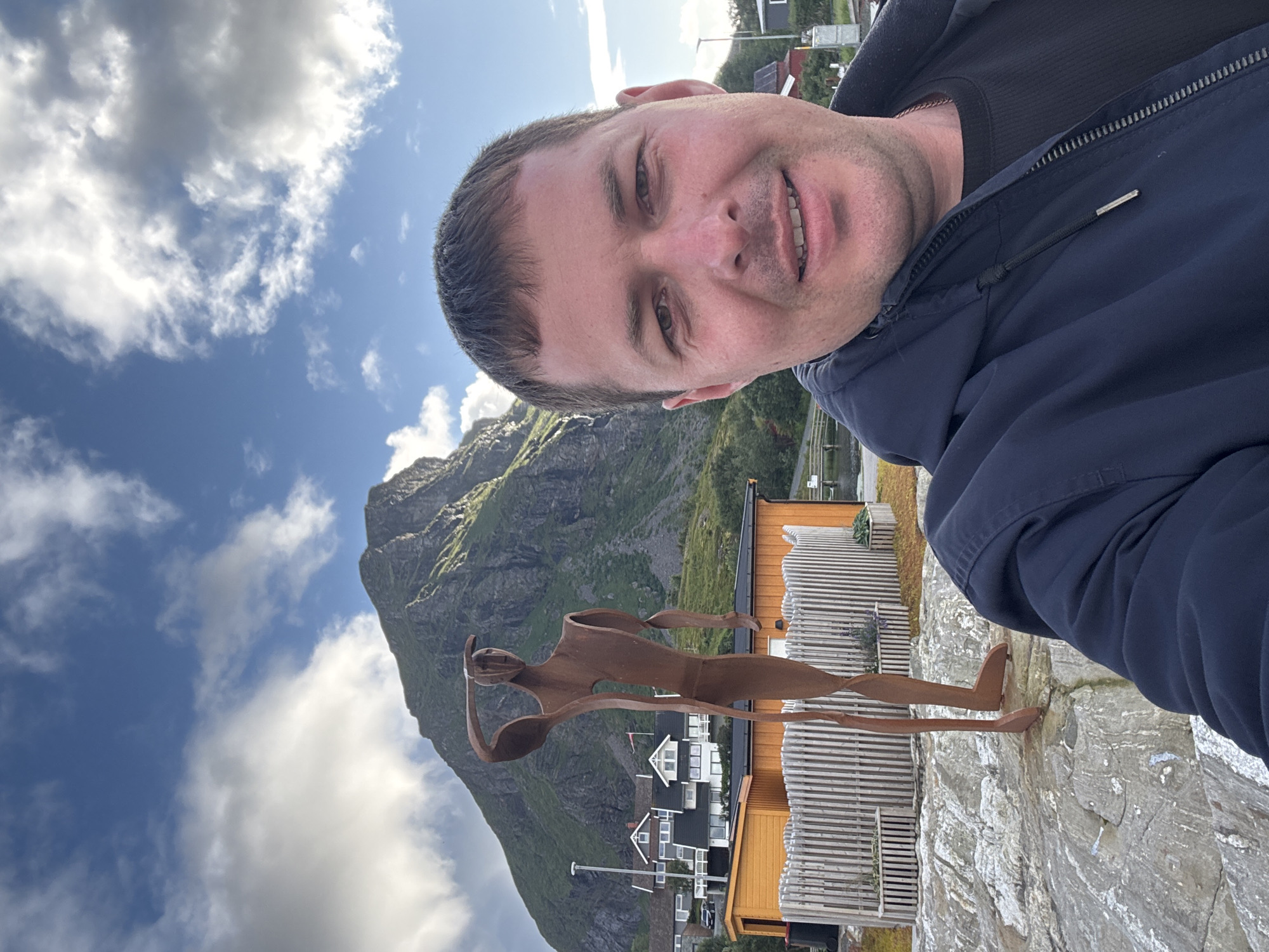 A man takes a selfie outdoors with a metal sculpture of a person in the background. The man is smiling and wearing a dark blue jacket and a black t-shirt. The sculpture is tall and slender with its right arm raised to its head. Behind the sculpture, there are orange buildings, a white fence, and a steep, green mountain under a blue sky with white clouds. This location is the city of Lovund, Norway.