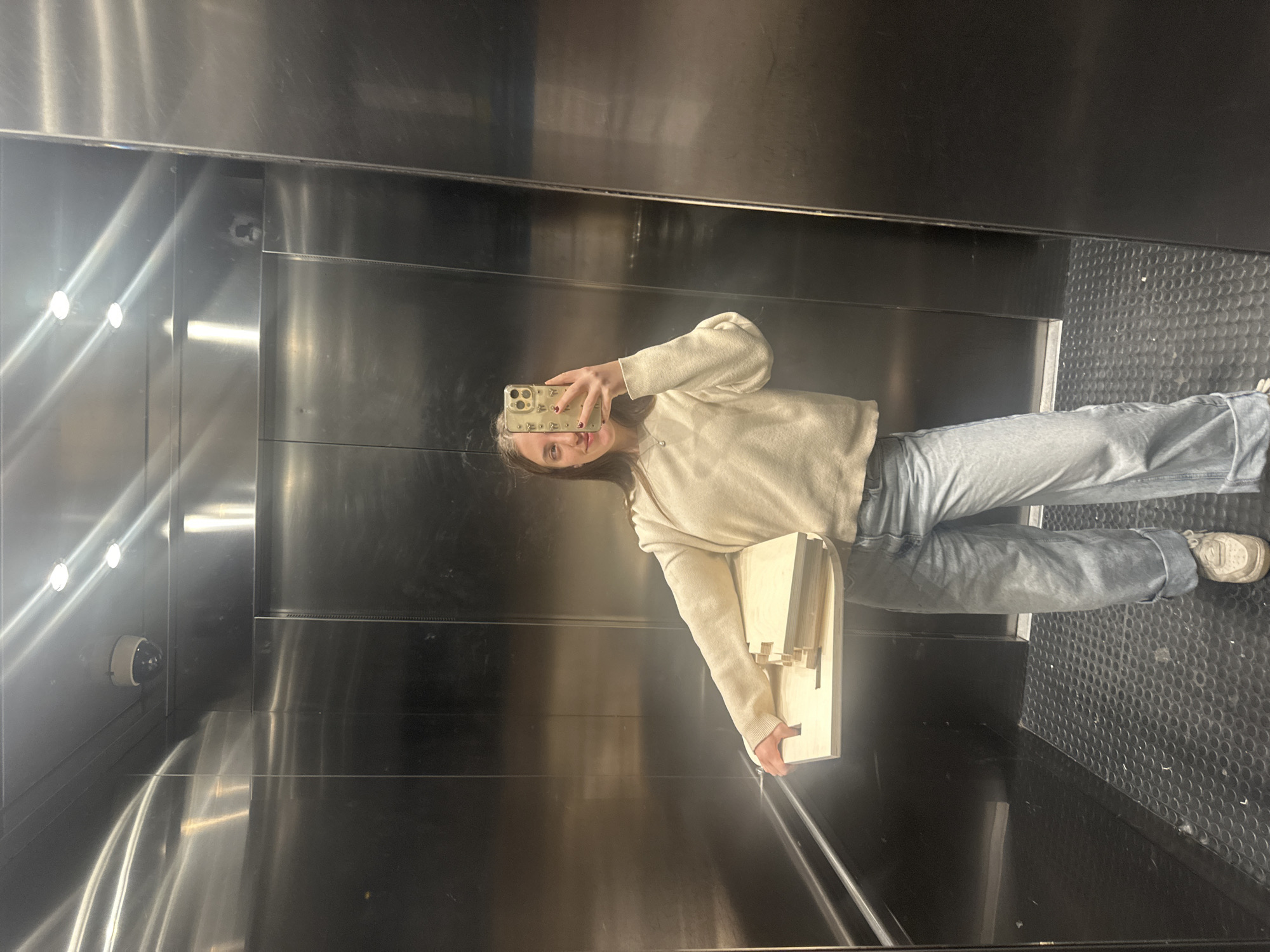 A young woman is taking a selfie in a stainless steel elevator. She is holding a model of a building and wearing jeans and a beige sweatshirt. The elevator has a surveillance camera in the upper left. The elevator's floor is black with a circular grid pattern. The location is unknown and there is no evidence in the image to indicate it is in Mexico City, Mexico.