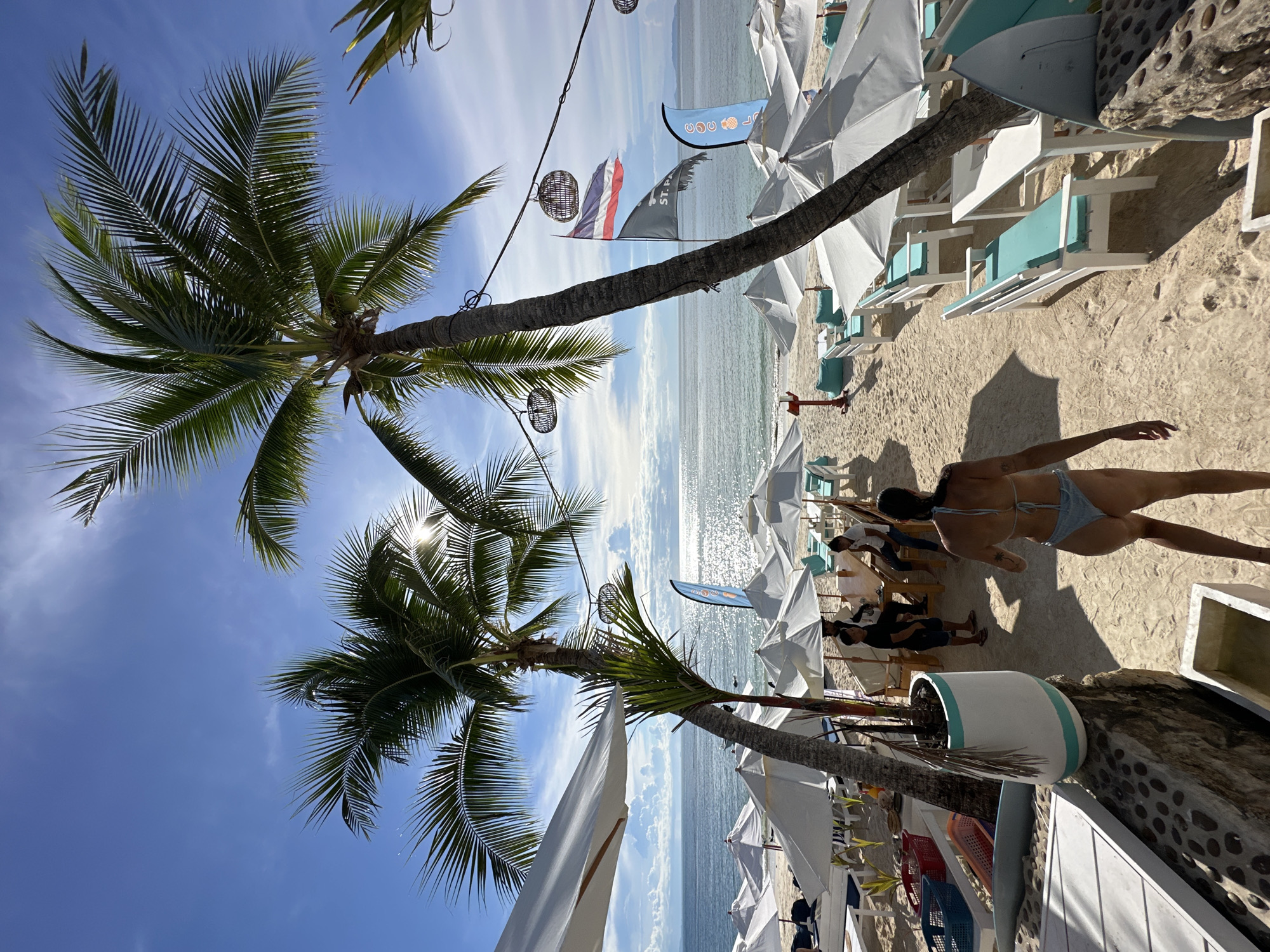 The image depicts a sunny beach scene at a resort in Koh Phangan, Thailand. In the foreground, a woman in a light blue bikini walks away from the camera towards a row of white tables and chairs with turquoise cushions, set up on the sand. Two palm trees, one in the foreground and one slightly behind it, frame the scene. Beyond the seating area, numerous white umbrellas are open on the beach, casting shadows on the sand. The ocean is visible in the background, with gentle waves and the sun glinting off the water. Several flags, including the Thai flag, are planted in the sand near the water. A few people are seated at tables further down the beach, and a couple of staff members are attending to them. The environment appears to be a resort or a beach club, with a relaxed and inviting atmosphere.