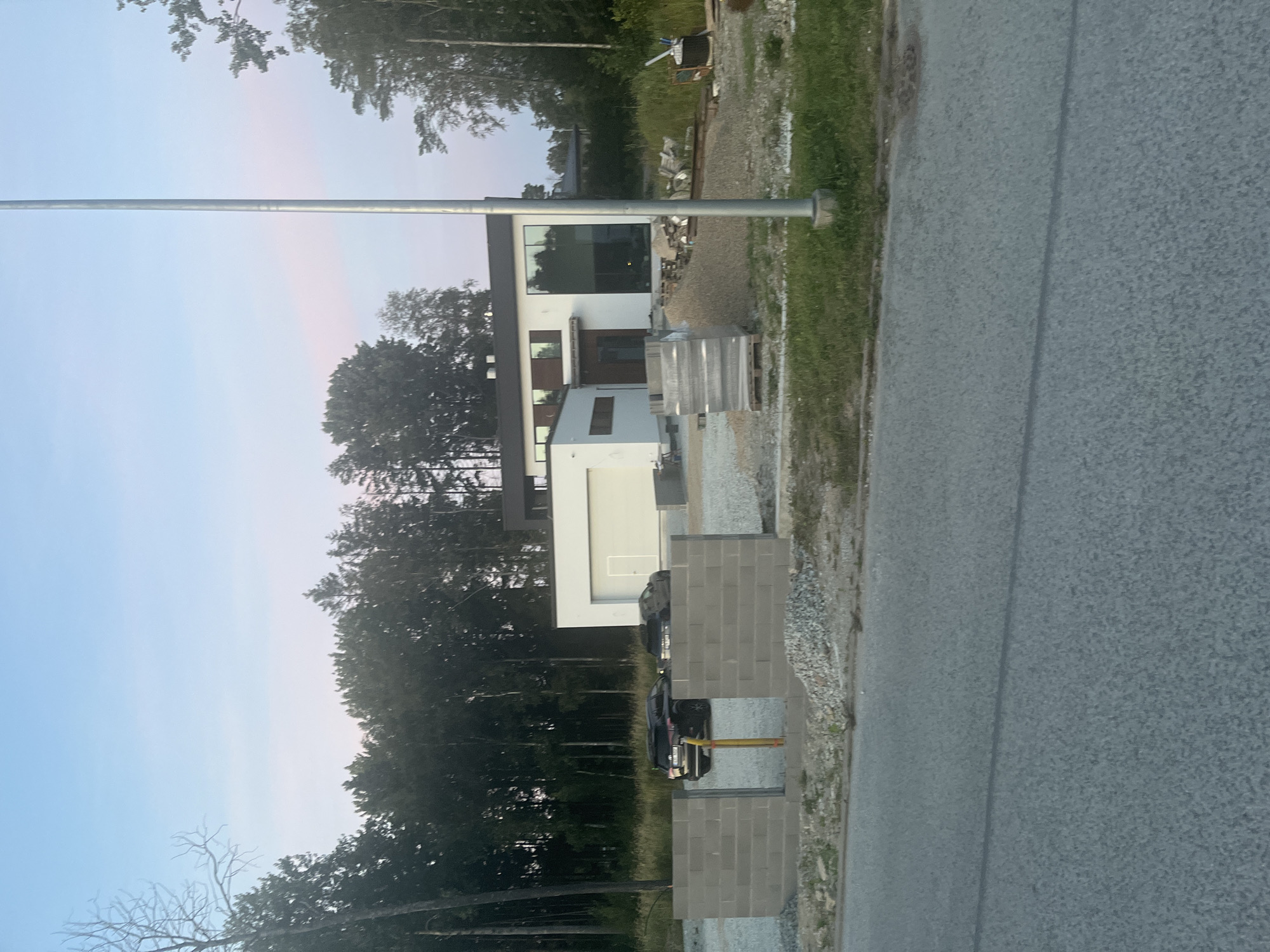 The image depicts a suburban residential construction site in Haabneeme alevik, Estonia, during dusk. In the background, a modern, multi-story house with large windows and a white facade is partially visible, surrounded by trees. In the foreground, a partially constructed gray brick wall is present, with a gap in the center. Several vehicles, including a black SUV and a dark sedan, are parked behind the wall. Construction materials, such as piles of gravel and stacked bricks, are scattered around the site. A tall, slender metal pole, likely a streetlamp, stands prominently in the middle of the frame. The sky is a gradient of pale blue and pink, indicative of twilight. The overall scene suggests an ongoing construction project in a residential area.