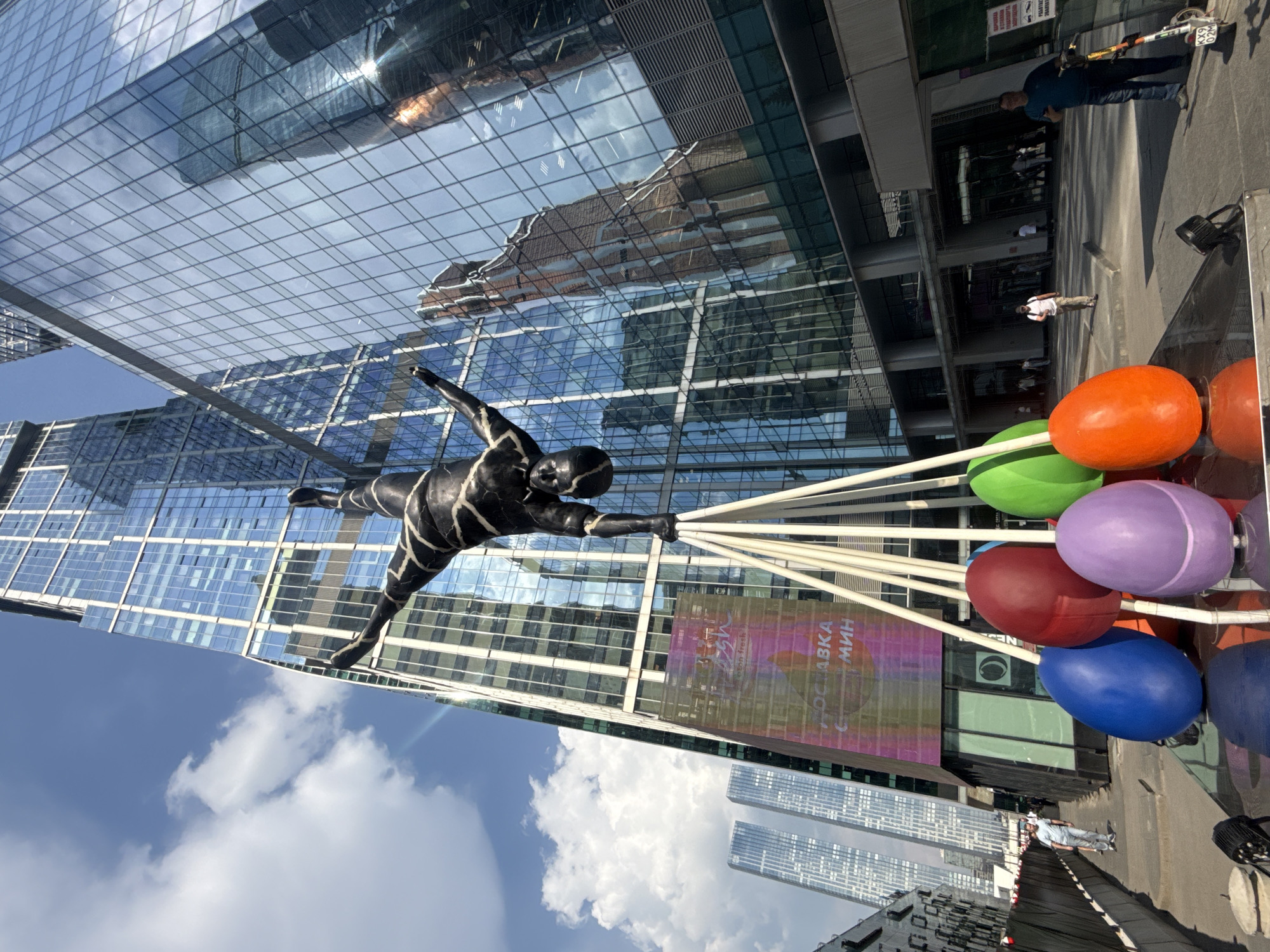 The image depicts a contemporary urban scene with a striking sculpture as the primary subject. The sculpture, a stylized human figure in a black and white striped outfit, appears to be in mid-flight, reaching out with one hand towards a cluster of large, colorful, egg-shaped balloons. These balloons, in vibrant shades of orange, green, purple, and blue, are suspended by white poles.
The setting is a modern city, characterized by towering glass skyscrapers that reflect the sky and surrounding buildings. The architecture suggests a business or financial district. A few people are visible in the background, including one man standing near a bicycle with a motor attachment, and others walking along a paved area. The sky is blue with scattered white clouds, indicating fair weather, likely during daylight hours.
The overall impression is one of dynamism and playful art integrated into a sleek, modern environment. The contrast between the fluid, dynamic lines of the sculpture and the rigid, geometric forms of the buildings creates a visually interesting composition. The reflections on the glass facades add depth and complexity to the scene. The presence of people suggests this is a public space, possibly a plaza or a street in a commercial area. There is some visible text on a banner in the background, appearing to be in Cyrillic script, partially obscured by the sculpture. The text seems to be "ОСТАВКА МИН" which translates to "Resignation of the Minister".