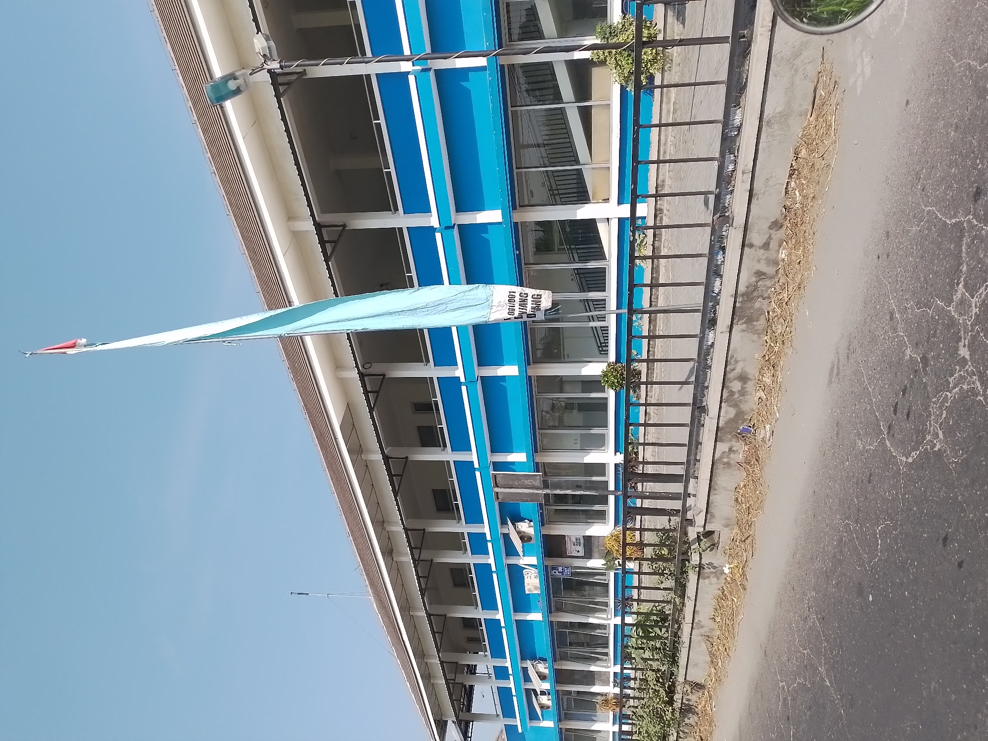 The image shows a two-story building with a blue and white facade, likely a government office or school, located in Ngancar, Indonesia. The building has a covered walkway on the second floor and many windows on the ground floor. A flag, partially obscured by a tall flagpole, flies in front of the building. The area in front of the building is lined with a black metal fence and a low concrete wall. Sparse vegetation and dry leaves are visible along the base of the wall and on the paved area in front of it. The road in the foreground appears cracked and weathered. A street lamp is visible attached to a pole near the building. The sky is clear and blue.