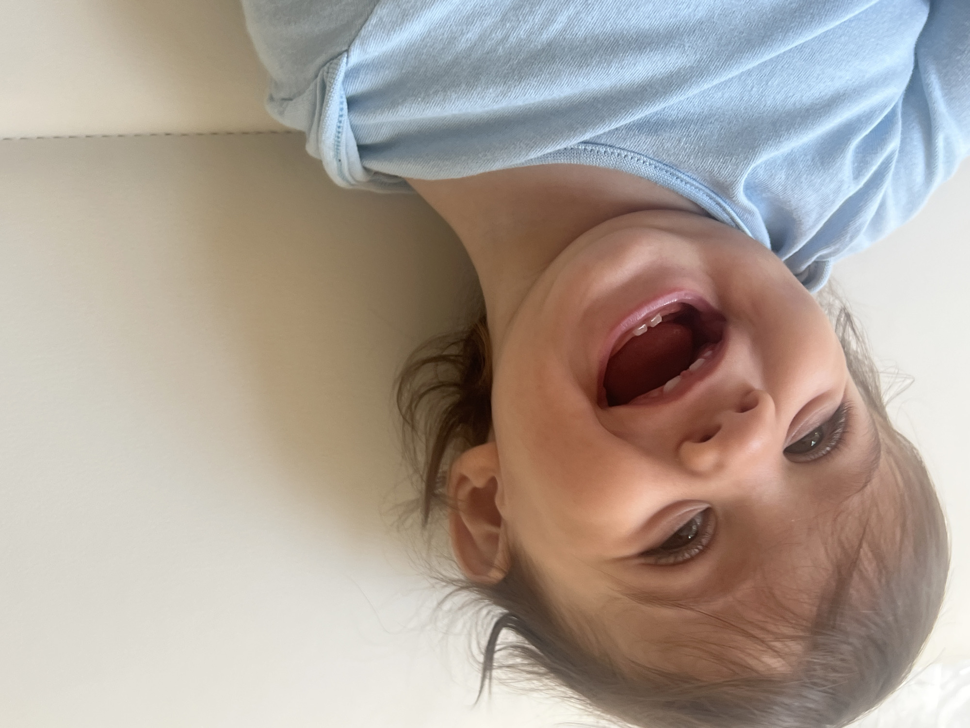A baby lies on its back on a white surface. The baby is wearing a light blue shirt and has brown hair and eyes. The baby is smiling with its mouth open, revealing a few teeth. The background is a plain white surface. The city of Elena, Bulgaria is not visible in the image.