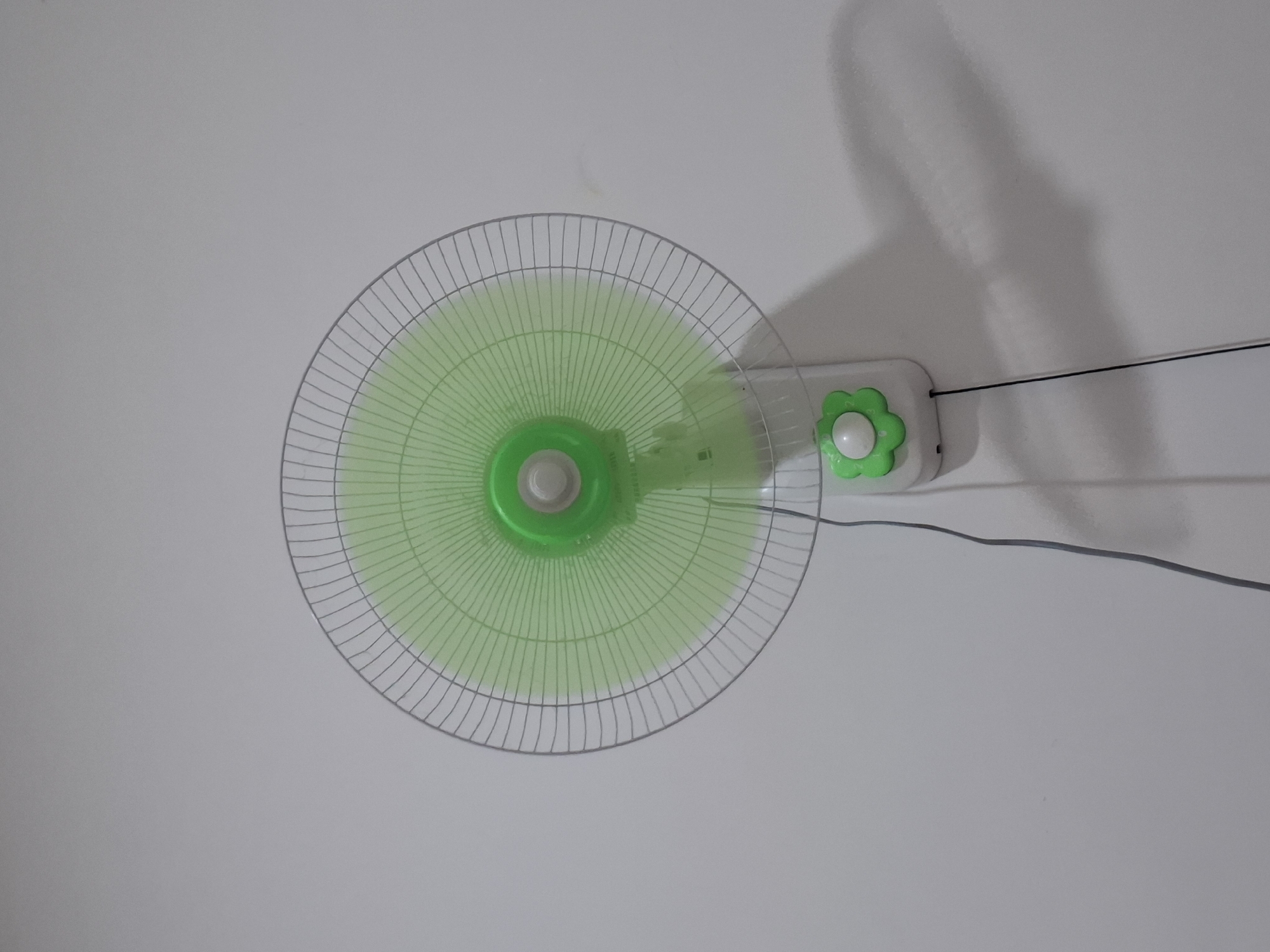 A close-up, eye-level, vertical shot shows a wall-mounted fan with a green oscillating fan blade and a silver metal grille. The fan is mounted on a white casing with a decorative green flower-shaped button that has numbers 1, 2, and 3 on it. A black wire hangs from the fan casing and trails down the wall, casting a shadow to the right. The wall is white and textured, with a faint shadow of the fan visible to the right of the fan itself. The fan appears to be in a residential setting, likely within a home in Hulaan, Indonesia.