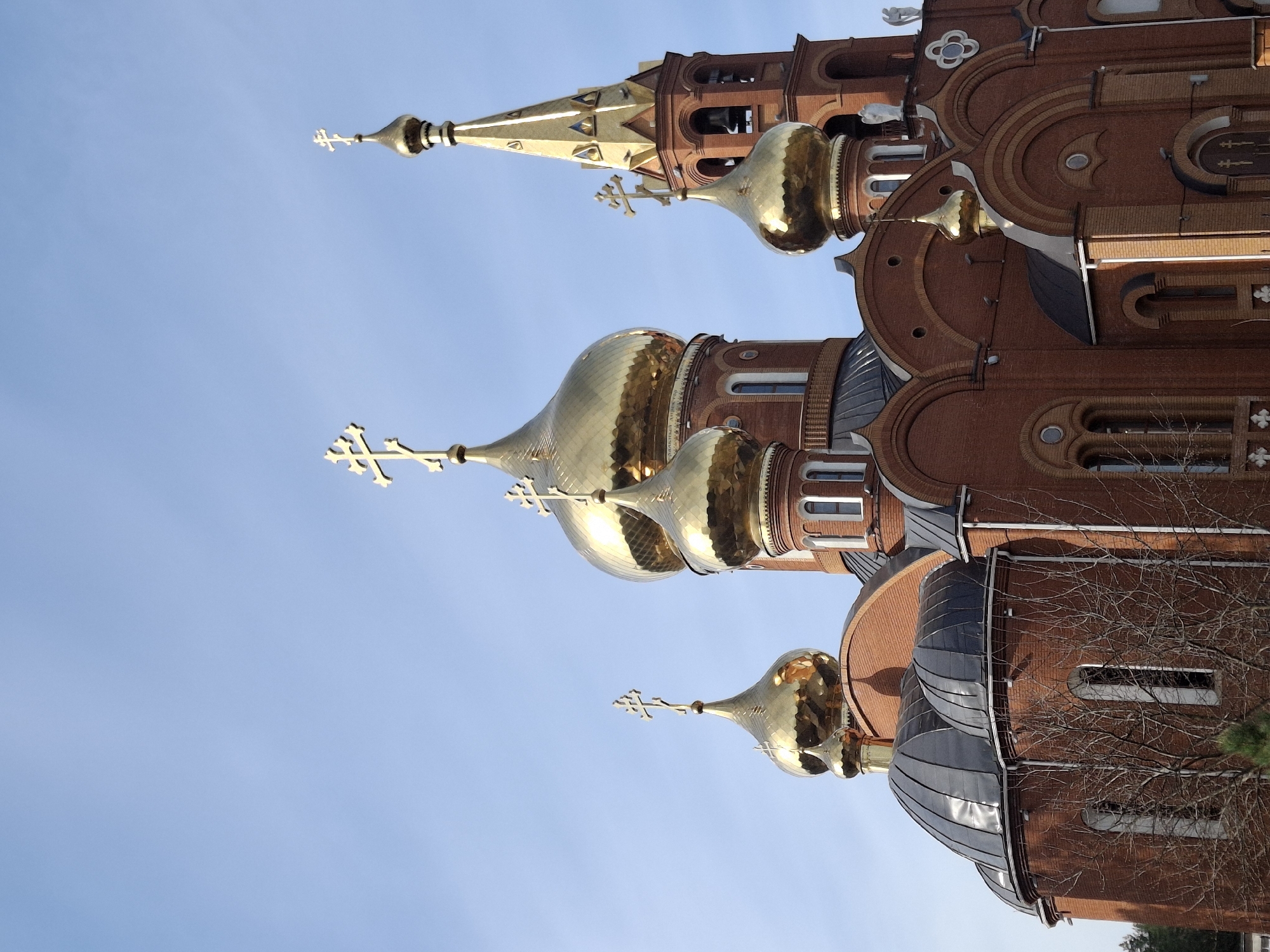 The image displays an imposing Eastern Orthodox church or cathedral, characterized by its reddish-brown brick construction and numerous golden, onion-shaped domes (cupolas), each topped with an ornate cross. The architectural style features intricate patterns in the brickwork, arched windows, and decorative elements along the facade. Darker, possibly metallic, roofs cover some sections of the building.
The scene is set outdoors under a clear, bright blue sky, indicating daytime and good weather. Sunlight reflects brightly off the gilded domes, highlighting their texture and form. No specific activity or event is depicted, as the image focuses solely on the building itself. Parts of bare tree branches are visible on the right side, suggesting the season might be late autumn or winter.
While some faint inscriptions or decorative patterns appear on the golden domes, they are not legible. The overall impression is one of grandeur and religious significance, typical of churches in the region. The image depicts a church located in Chaikovsky, Russia.