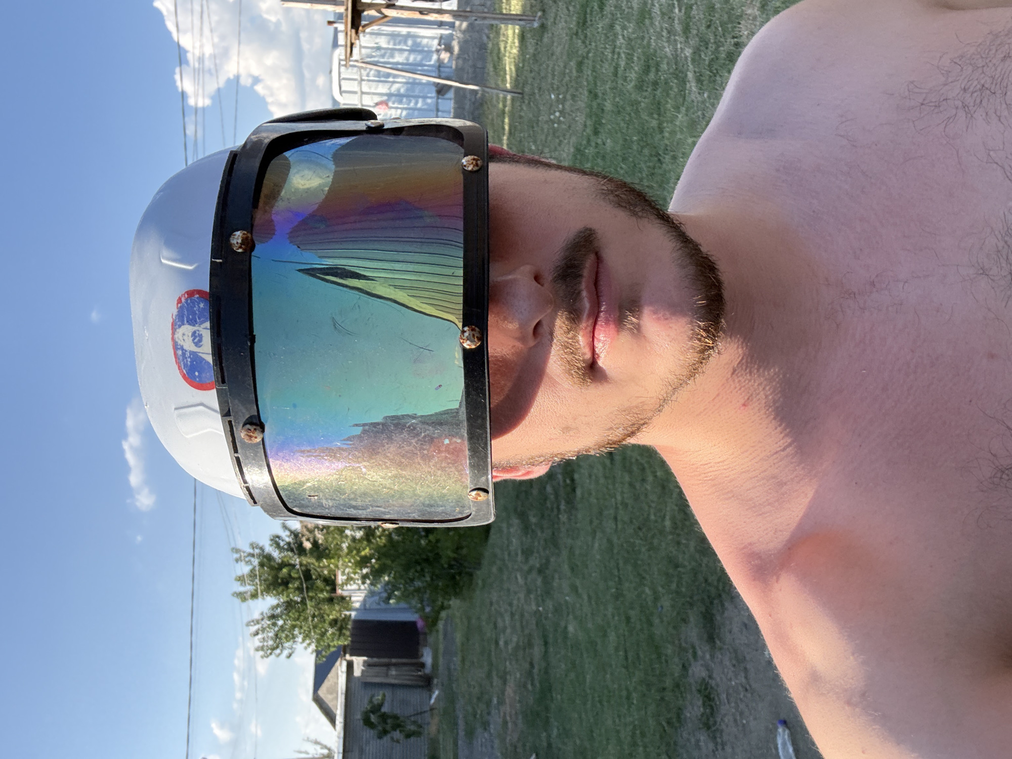 A young adult male stands outdoors, facing the camera. He is shirtless and has facial hair. He is wearing a white hard hat with a multi-colored, reflective visor that covers his eyes and forehead. The visor has visible scratches and smudges. The background suggests a rural or suburban setting with a grassy yard, a wooden fence, and a building in the distance. The sky is blue with scattered clouds. There is no indication of the location being Tbilisi, Georgia.