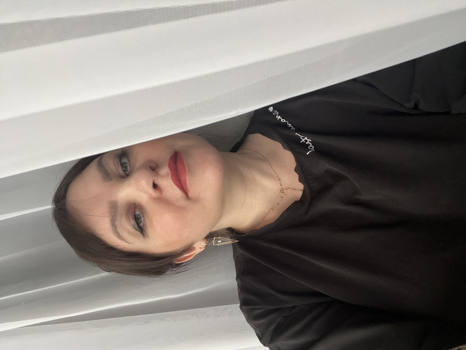 Noviy, Russia: The scene depicts a female subject in an indoor environment. She has light skin, dark brown hair, and is wearing red lipstick and eyeliner. Her gaze is directed towards the camera. She is wearing a loose-fitting black t-shirt with a ruffled neckline and white embroidered text on the right chest area. A gold-colored lariat-style necklace with a circular accent is visible around her neck. On her left ear, she wears a gold-colored, upward-pointing triangular earring with a clear central element, along with a small gold-colored hoop earring. The background consists primarily of vertically pleated, translucent white fabric or curtains, which partially obscure the subject's right side and a portion of the left background. A darker, vertical linear element is faintly visible behind the curtain on the left.