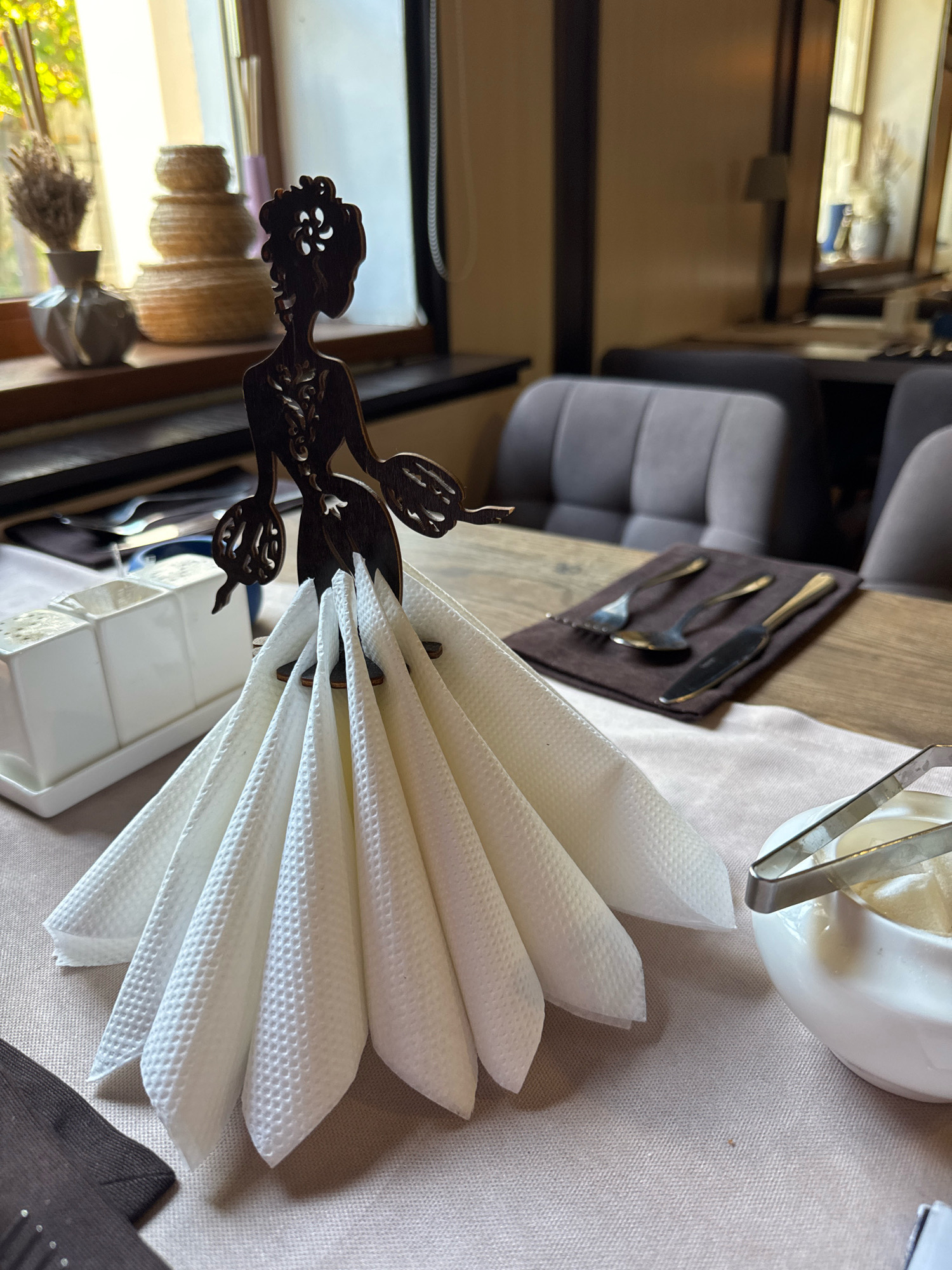 The image depicts a close-up of a table setting in what appears to be a restaurant in Staraya Russa, Russia. A decorative wooden cutout of a woman in a formal gown is used as a napkin holder, with white, textured napkins fanned out to create the "skirt" of the dress. The figurine is dark brown and intricately detailed, with a floral design on its bodice and headpiece.
In the background, a window shows greenery outside, suggesting a daytime scene. On the windowsill, there are stacked woven baskets and a vase with dried flowers. The table itself is set with a light pinkish tablecloth and a wooden surface. To the left of the napkin holder is a white rectangular dish with compartments, possibly for condiments or sugar. To the right, a small white bowl with sugar cubes and silver tongs is visible. Further back on the table, a dark brown cloth holds a set of silverware, including at least two spoons and a knife. In the blurred background, upholstered chairs are visible, indicating a dining area. The overall impression is one of a quaint and thoughtfully decorated establishment.