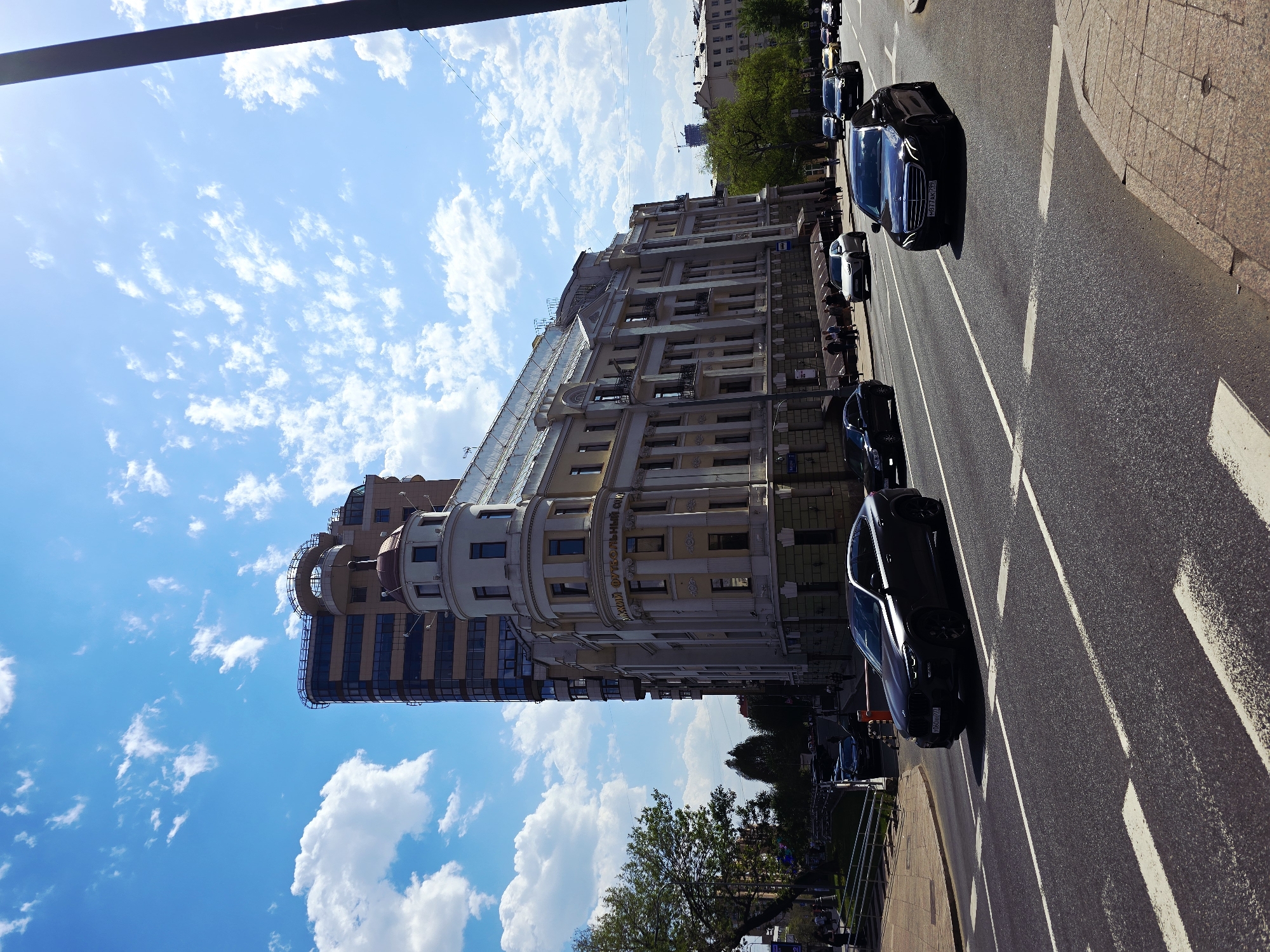 The image captures a street scene in Moscow, Russia, prominently featuring a building complex juxtaposed against a blue sky filled with cumulus clouds. The building has a historical facade with decorative elements and a modern glass-clad tower at one end. Vehicles are traveling along a multi-lane road marked with lane dividers. The presence of trees lines the street near a sidewalk. The buildings have visible signage, including the name of the sports complex.