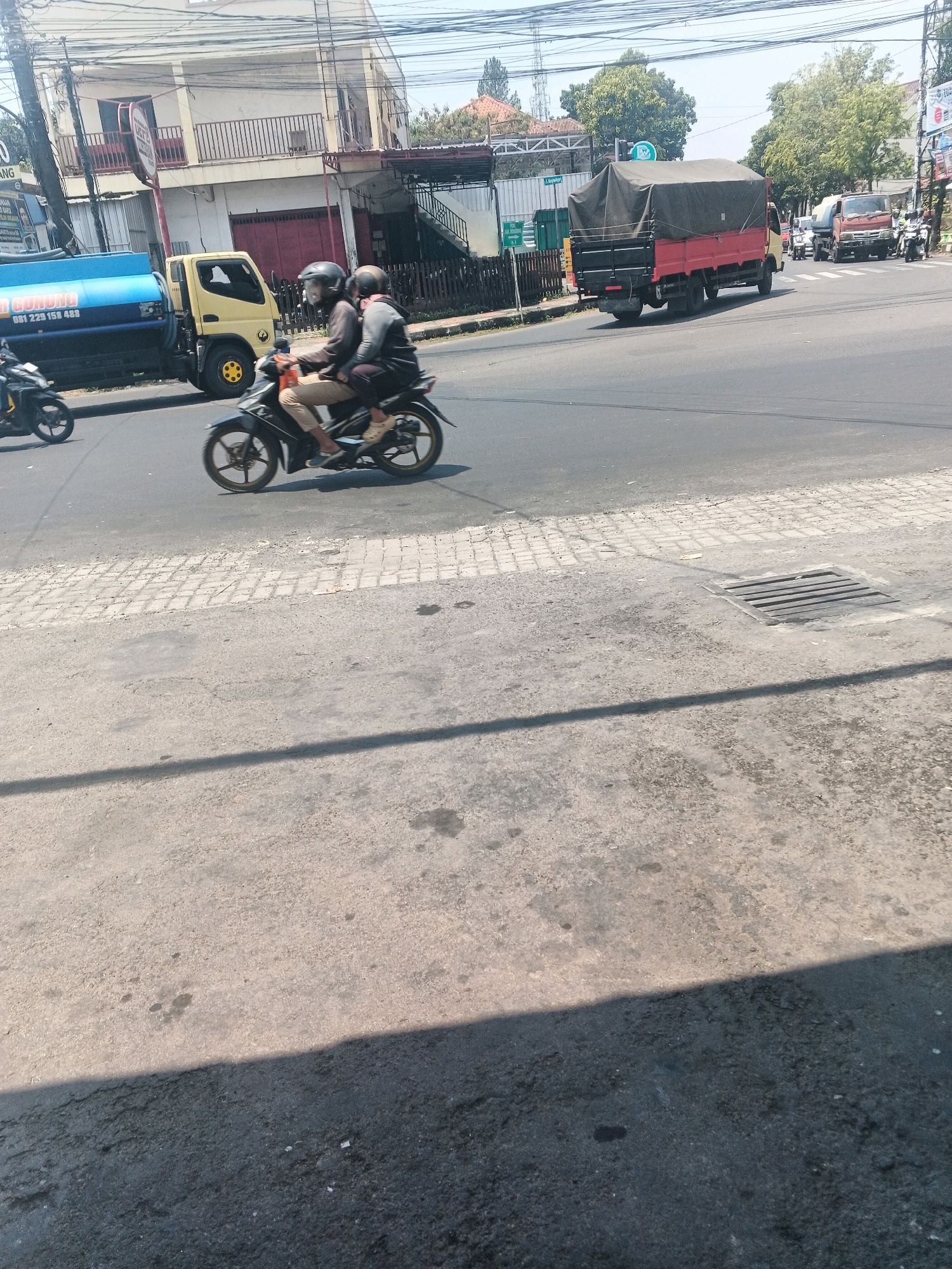 In Ungaran, Indonesia, on a bright, sunny day, a two-person motorcycle passes by a yellow water tank truck. The motorcycle is traveling on a paved road alongside a sidewalk made of interlocking bricks. In the background, a red truck with a covered cargo bed is parked, and further down the road, another truck and several motorcycles are visible. The scene is set against a backdrop of buildings and trees, with power lines overhead.