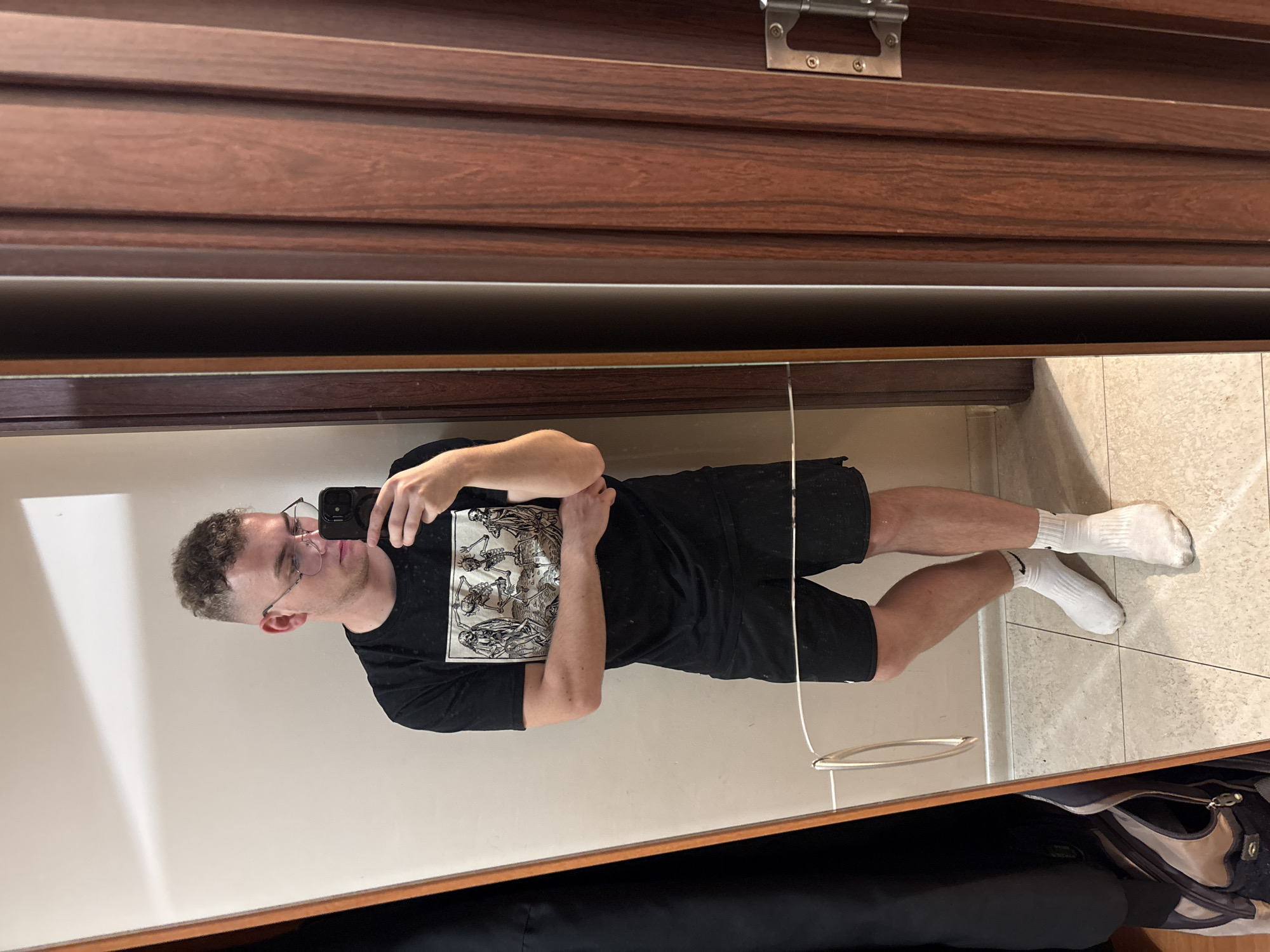 This mirror selfie, taken indoors, features a young man in Plovdiv, Bulgaria. He appears to be in his 20s or 30s, with short, curly brown hair and glasses, holding a dark-colored smartphone to capture his reflection.
The man is dressed casually in a black t-shirt adorned with a prominent white graphic print depicting skeletons and macabre art. He pairs this with black shorts and white crew socks; a Nike swoosh logo is visible on his right sock. His expression is neutral, focused on the act of taking the photo.
The indoor setting shows a light-colored, possibly beige, wall reflected behind the man, while the floor is tiled with light-colored squares and visible grout lines. Above the mirror, dark brown wooden paneling with horizontal grooves, suggestive of a wardrobe or cabinet door, spans the top of the frame. A silver metal hinge or latch is fixed to this paneling. A thin white cord or wire hangs vertically in front of the mirror, near the man's legs, and a silver oval-shaped handle is also reflected on the tiled floor. Below the mirror's frame, a dark, recessed area contains a partially visible light-colored bag or object, likely part of a storage unit.