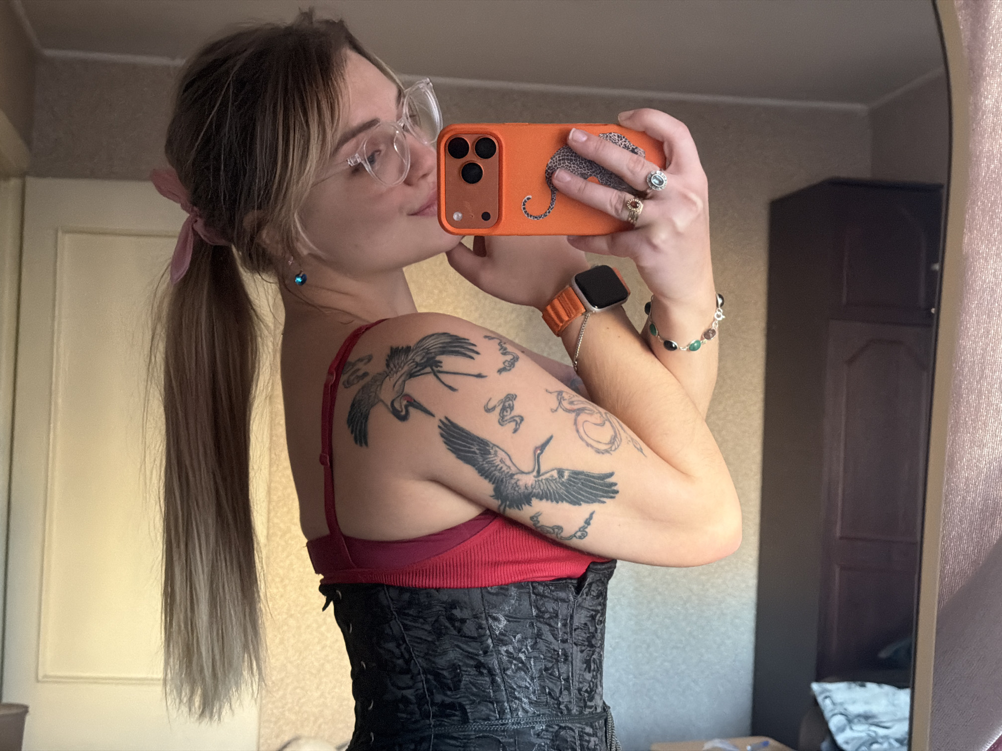 A fair-skinned woman is seen in a mirror selfie indoors. She has blonde hair styled in pigtails with a pink ribbon and wears glasses. She holds an orange-cased phone with a leopard print design. Her attire includes a red top under a black corset, and she has several tattoos on her left arm featuring crane-like birds. She wears an orange smartwatch and a bracelet with gemstones. A ring is visible on her finger. The background shows a beige wall with white trim to the left, and a dark brown cabinet to the right. The location is in an unspecified interior, but information on the city of Naberezhnye Chelny, Russia is not visibly supported by the image.
