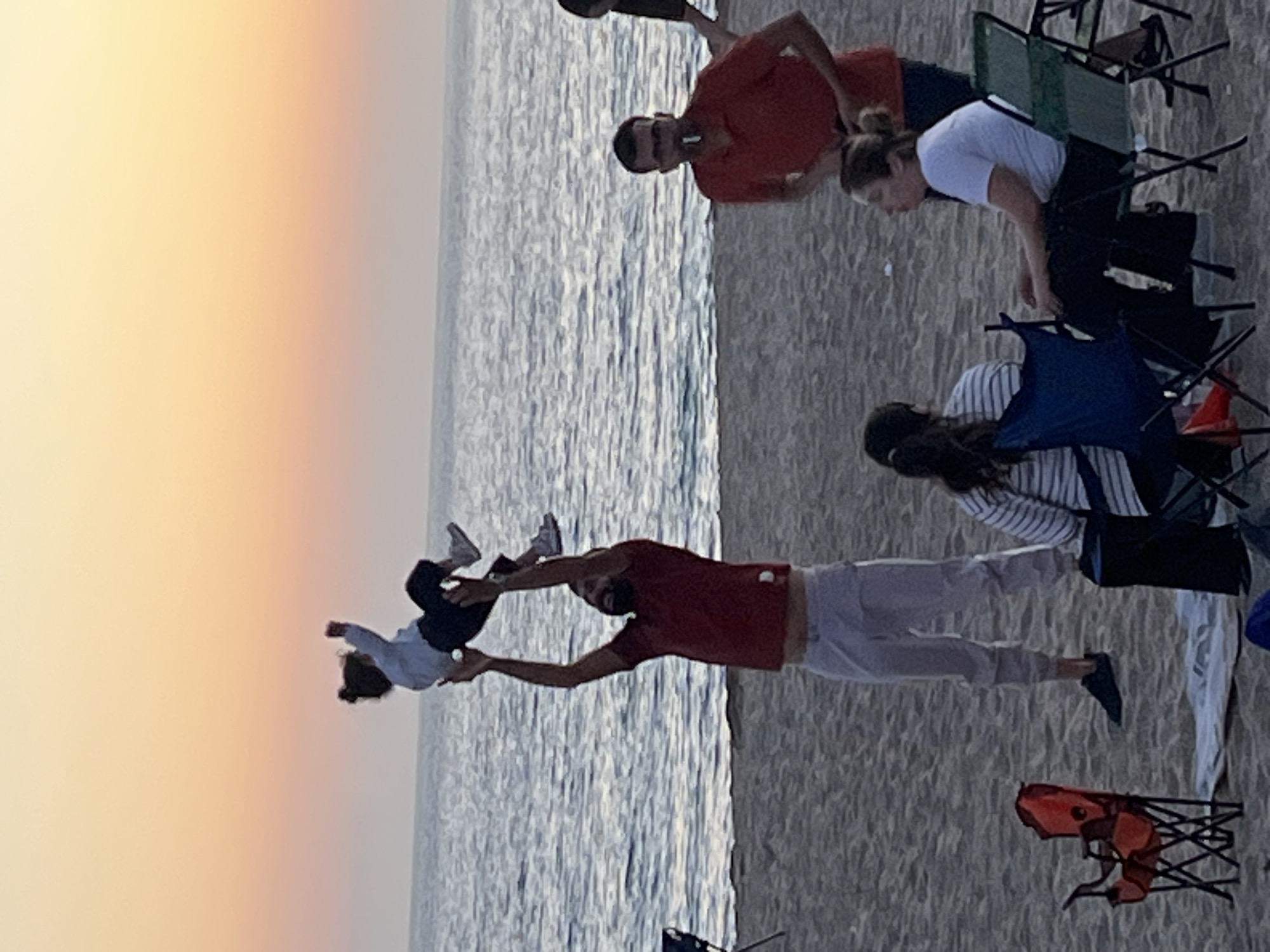 The image depicts a group of people on a beach. The location is likely in Alanya, Turkey, based on the presence of the ocean and the sunset in the background. A man in a red shirt is holding a child in the air, with the child appearing to be flying. Several other people are present, some seated in folding chairs and others standing nearby. The overall environment suggests a relaxed, recreational setting. The beach appears sandy, with the ocean water visible in the background. The sky shows colors of the setting sun.