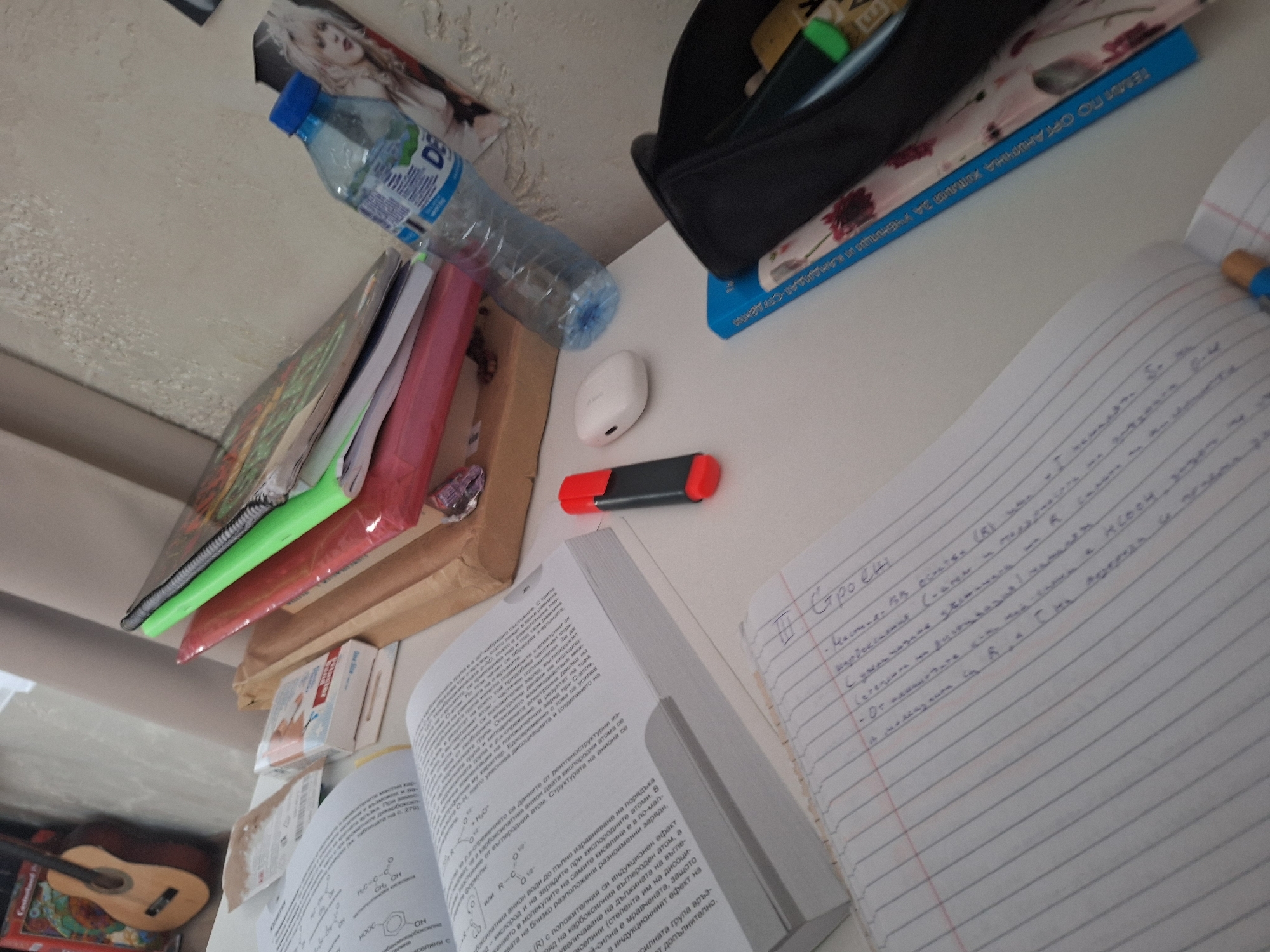 The image depicts a cluttered desk or table, possibly used for studying, with various objects scattered across its surface. There is a pile of books and folders to the left, topped with a transparent folder with green binding. A bottle of water is lying horizontally on top of the books. Next to the books, there are some small boxes or packages of medication. A pair of white wireless earbuds and a black and red marker are positioned near the center of the table. An open textbook, written in Bulgarian, and a notebook filled with handwritten notes are in the lower right corner of the frame. Above the textbook and notebook, there is a black pencil case with a blue book underneath. In the lower left, there is a partial view of a small guitar on a decorative case. The overall scene suggests a study environment with a focus on chemistry-related topics. The image does not provide any location in the city of Panagyurishte, Bulgaria.