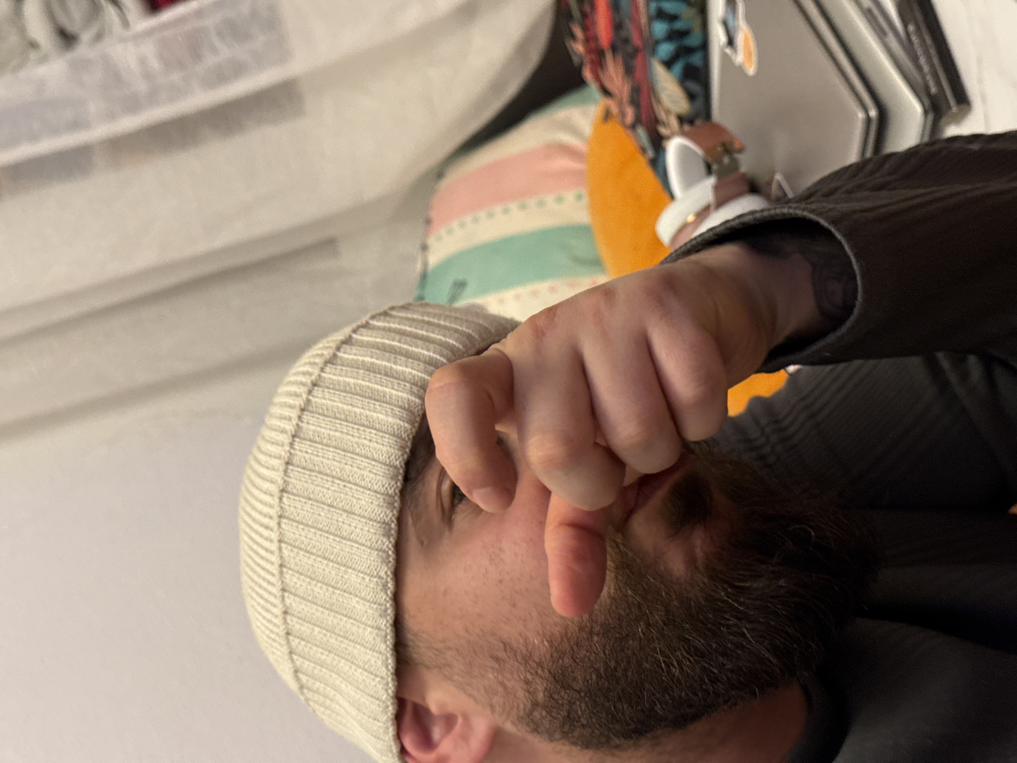 The image displays a close-up, chest-up shot of a male individual indoors. The subject possesses a dark brown beard and fair skin, and wears a light beige ribbed knit cap pulled down over his forehead. He is dressed in a dark grey long-sleeved shirt or jacket. His right hand is raised, with the index finger extended directly towards the viewer, while the remaining fingers are partially curled. A white wristwatch with a light pink band is visible on his left wrist, along with a partial circular tattoo on his left forearm. He looks directly forward, with his mouth slightly ajar. The background to the left features a plain, light-colored wall. To the right, a sheer, light-colored curtain hangs, partially obscuring a darker area, possibly a window. Behind the individual, a striped pillow featuring light blue, pink, and white vertical bands is visible, alongside an orange object. A stack of silver-colored electronic devices, consistent with laptops, is present on a surface to the far right, with a sticker affixed to the top device. The scene represents an indoor residential setting. No visual cues within the image allow for the identification of a specific location within Strasbourg, France.