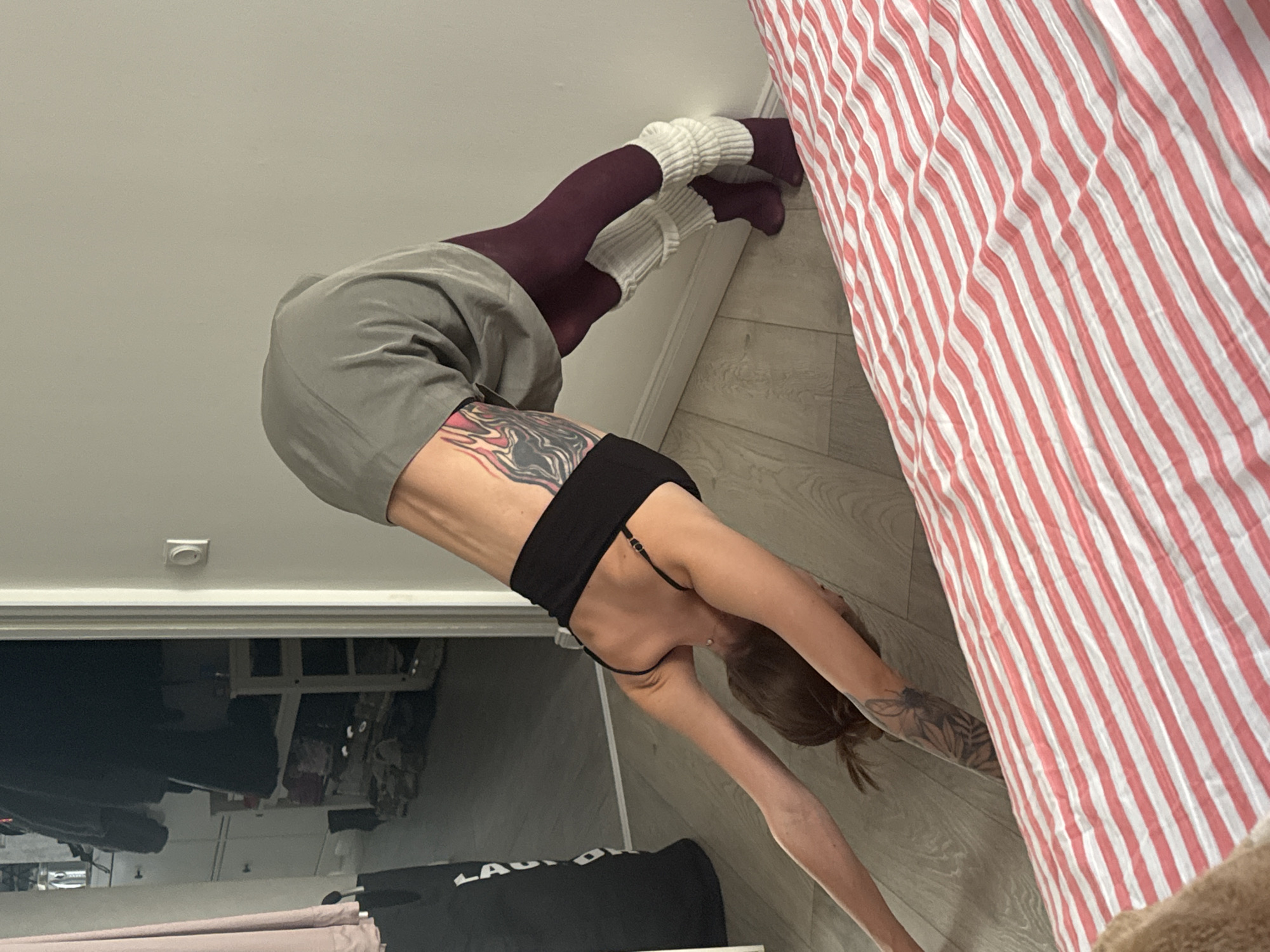 A woman is performing a downward-facing dog yoga pose against a light-colored wall. She is wearing a black tank top, a grey skirt, dark red tights, and white leg warmers. She has tattoos on her side and arm. The floor is wooden. In the background, a doorway leads to a room with a shoe rack and clothing. To the right, a bed with a pink and white striped comforter is visible. This scene is indoors, likely in a residential setting. There is no indication of location in Moscow, Russia.