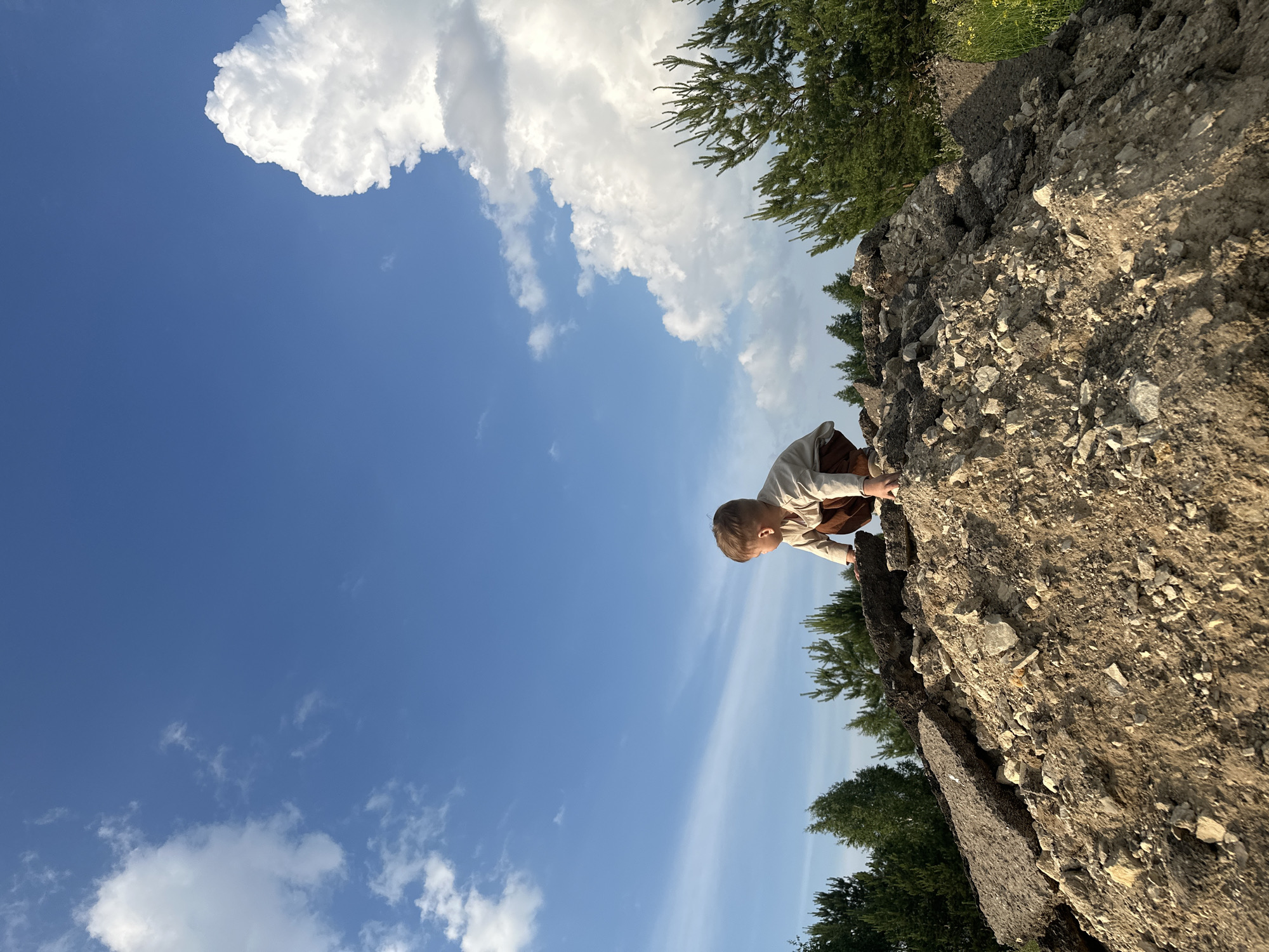 A young child, seen from behind, is crouched on a pile of rubble, possibly asphalt and dirt, under a bright blue sky with scattered white clouds. The child is wearing a light-colored long-sleeved shirt and brown pants. Behind the rubble pile is a line of green pine trees. The location is described as Estachi, Russia. The lighting suggests it is daytime, possibly late afternoon due to the warm tones in the rubble.