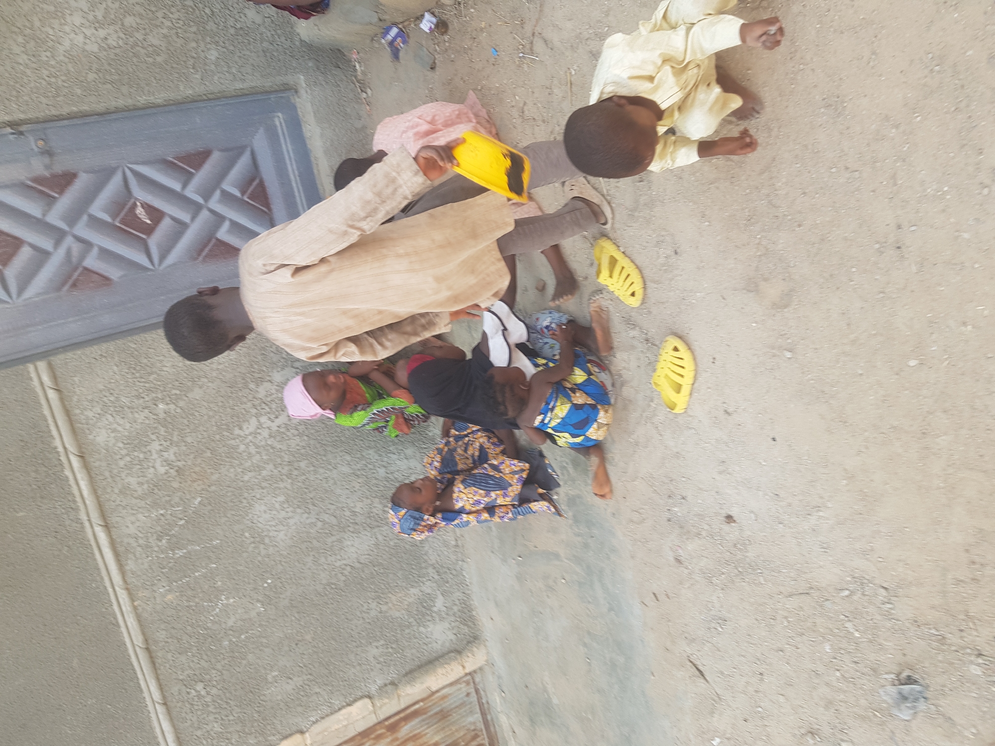 In Dutsinma B, Nigeria, a group of children are gathered on a dirt and concrete surface next to a building with a grey door. One child, wearing a light brown outfit, holds a yellow object. Another child is in a light yellow garment. Additional children are wearing colorful patterned fabrics, with some lying on the ground. One child wears yellow shoes. The area appears to be an outdoor space.