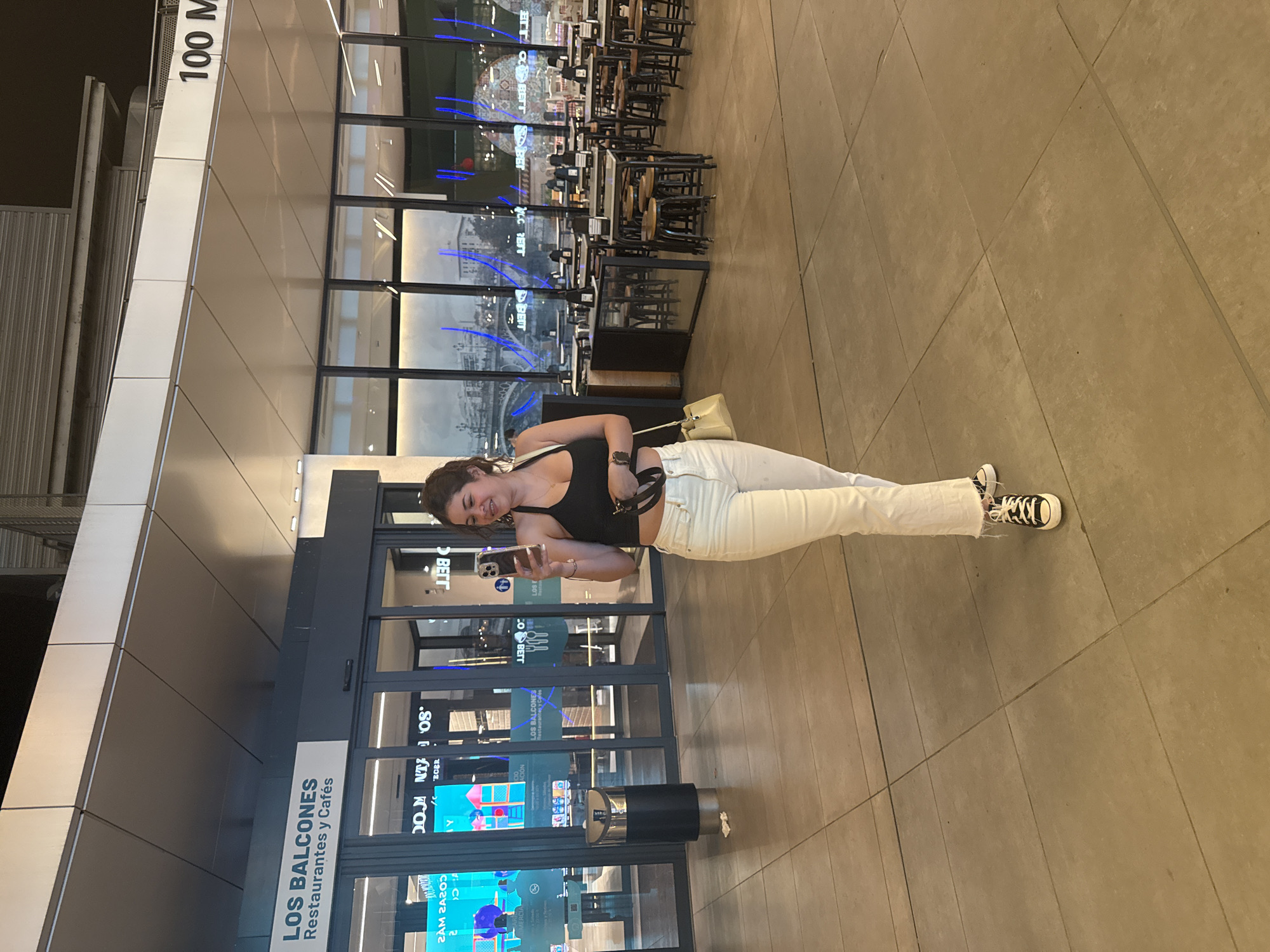 In Alcorcón, Spain, a young woman stands by the entrance of a restaurant and cafe called "Los Balcones." She is dressed in a black tank top and white pants, with black and white high-top sneakers. A light-colored purse hangs from her shoulder. The woman is holding a smartphone in her hand, as if taking a selfie or checking it. She appears to be smiling, looking down at her phone with a relaxed expression.
Behind her, through the large glass doors, several tables and chairs are visible within the restaurant, with some stacked neatly. Blue neon lights are reflected on the windows, suggesting the interior lighting or decor. The floor is tiled in a tan, grid pattern. To the left of the entrance, a sign displays "100 M" vertically on a white panel. The overall scene suggests an indoor public space, possibly a shopping mall or a transit hub, during the day or early evening, given the interior lighting. The atmosphere seems casual and modern.
