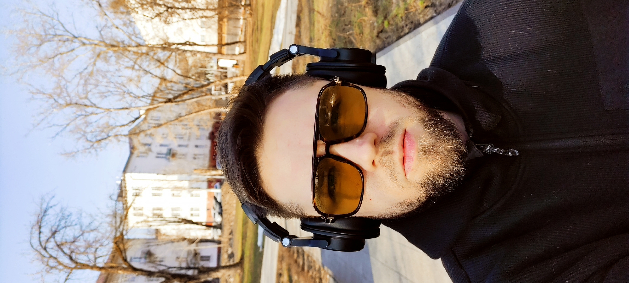 A man is taking a selfie outdoors. He wears sunglasses and black headphones, with a black jacket zipped up near the collar. The background shows trees and buildings, indicative of an urban setting. It is Fryazino, Russia.