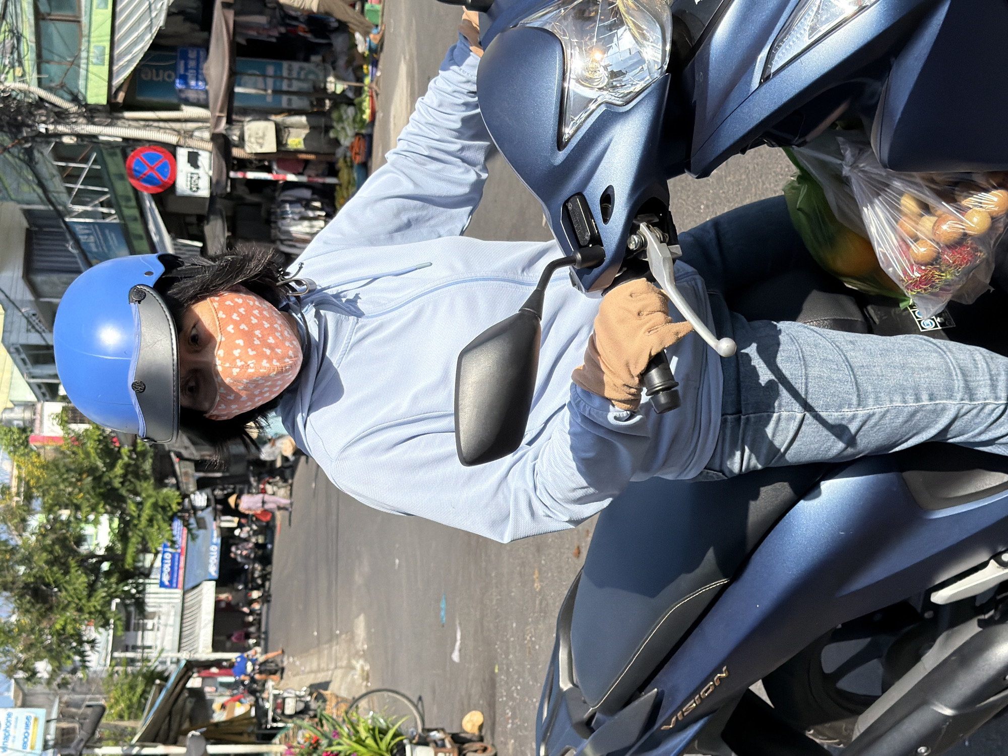 In a sunlit, bustling urban street scene in Nha Trang, Vietnam, a person wearing a blue helmet and a light blue hooded jacket rides a dark blue scooter. The individual is also wearing a light pink face mask with a white heart pattern and light-wash denim jeans. They are wearing tan gloves and their hands are on the scooter's handlebars. A bag of fruit, including what appears to be rambutan, is visible in front of the rider on the scooter. The background shows typical Vietnamese street life, with shops, buildings, power lines, other scooters, and pedestrians. The weather appears to be sunny.