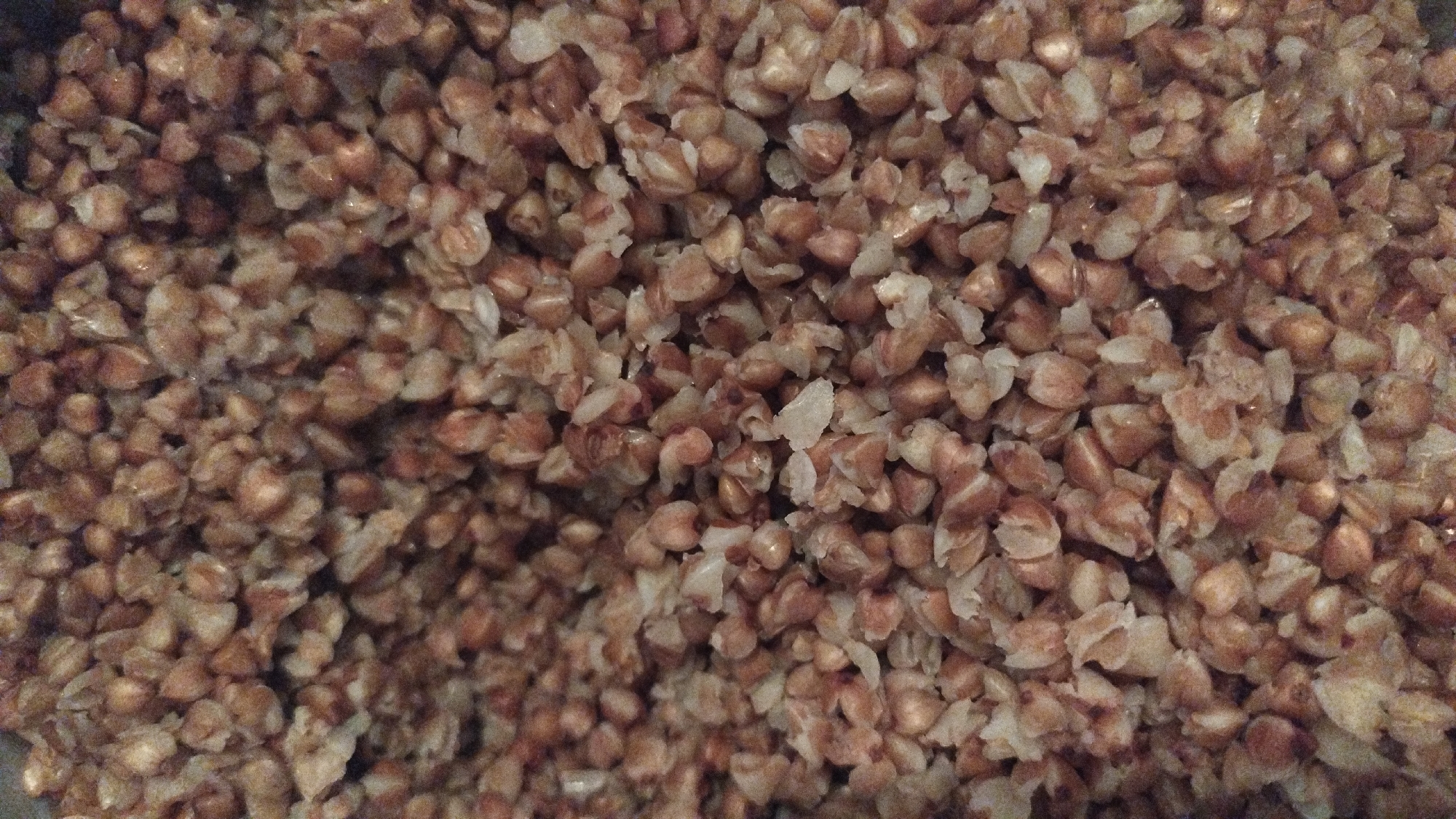 This is a close-up, overhead shot of cooked buckwheat groats. The grains are a light brown to reddish-brown color, with some lighter, almost white pieces visible, likely representing the inner kernel or partially cooked parts. The groats appear to be slightly moist, with some clumping together, suggesting they have been recently cooked or prepared.
The image is a still life composition focusing entirely on the food item. There are no people, animals, or discernible background elements. The lighting is diffused, creating soft shadows and highlighting the texture of the grains. The overall impression is one of simple, wholesome food, possibly being prepared for a meal. There is no visible text in the image. The setting is unknown, and the time of day and weather conditions are not discernible from the media.