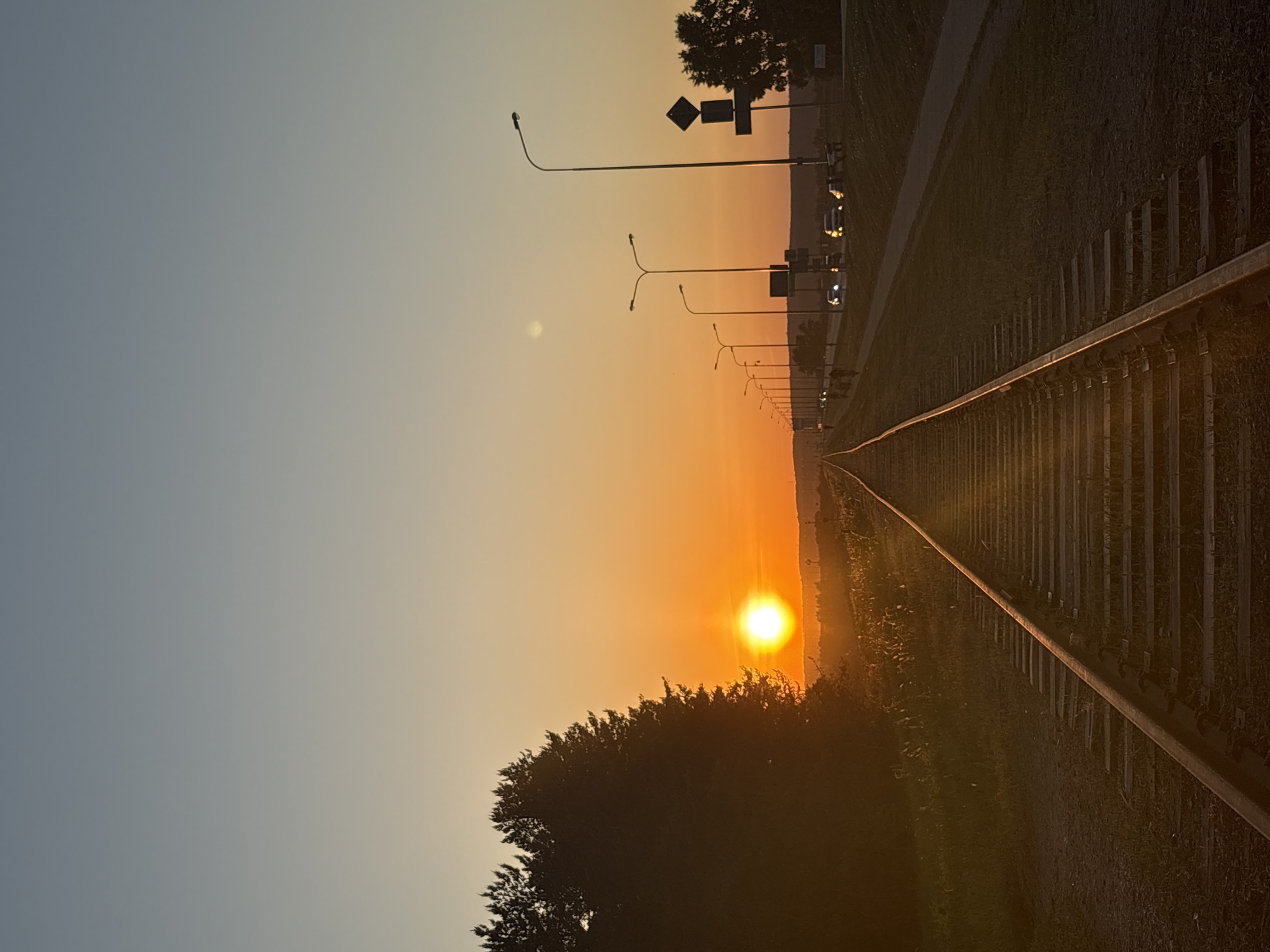 A sunset photo taken in Chaikovsky, Russia, shows a railway track receding into the distance where the sun is setting. The track has wooden supports, and on the right side of the frame is a grassy area. A line of streetlights and a road with cars are visible in the distance. On the left is a patch of trees. The sky is a gradient of light blue to orange, illuminated by the sun.