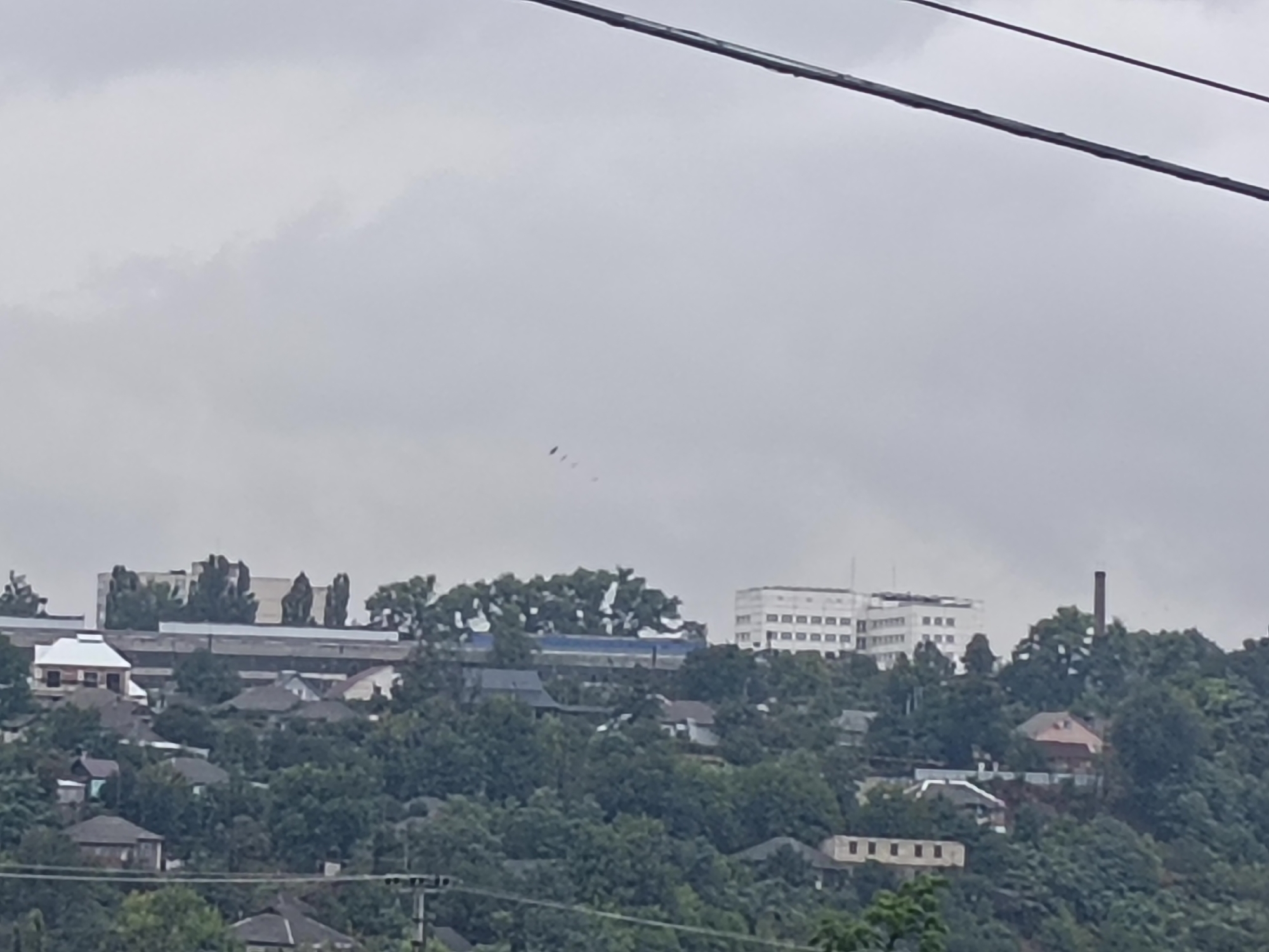 The image shows a hazy, overcast sky over a hillside populated with buildings and dense foliage. In the foreground, power lines are visible stretched across the frame. The midground reveals a cluster of houses, some with dark roofs and others with lighter ones, interspersed with trees. Further up the hill, larger buildings are apparent, including a multi-story white structure that appears to be a modern institutional building, possibly a hospital or a school, and a reddish-brown brick industrial chimney. The overall impression is of a residential and industrial area situated on a natural incline, under a somber sky. The scene is likely a panoramic view of a section of Rybnitsa, Moldova, given the distinctive architectural style of the larger buildings and the presence of industrial elements common in the city.