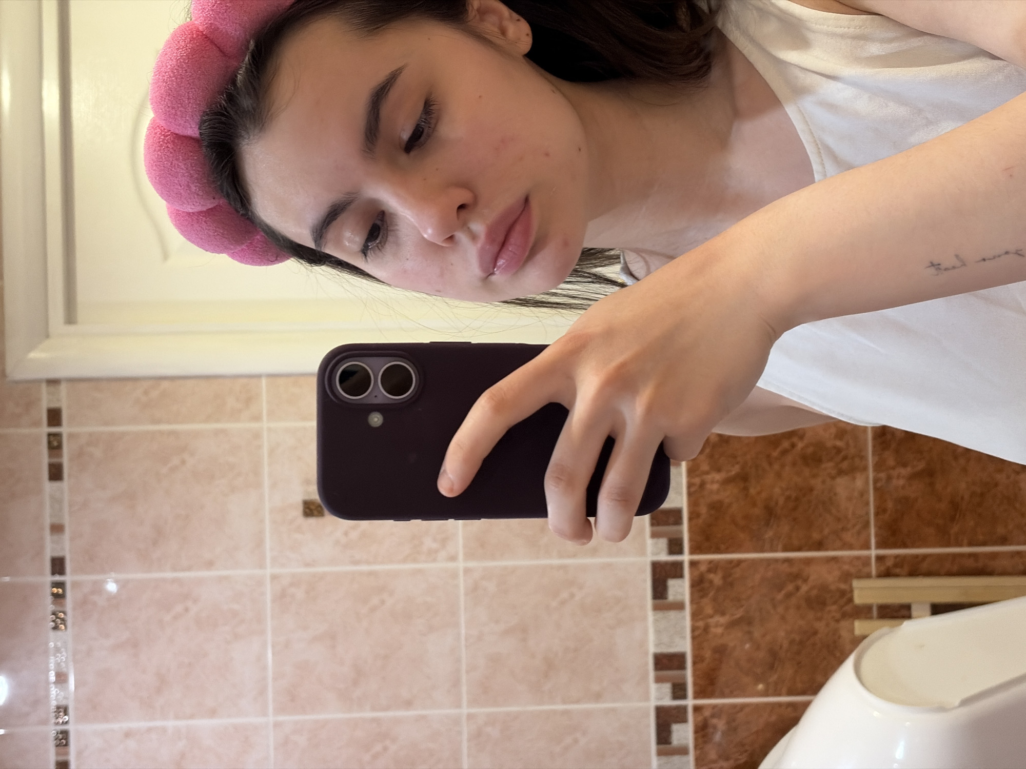 A selfie taken in a bathroom with light beige and brown tiled walls. A woman with dark hair wearing a pink headband is holding a dark-colored smartphone and taking the picture. She is wearing a white top. Part of a toilet is visible in the lower right corner. The location is Chaikovsky, Russia.