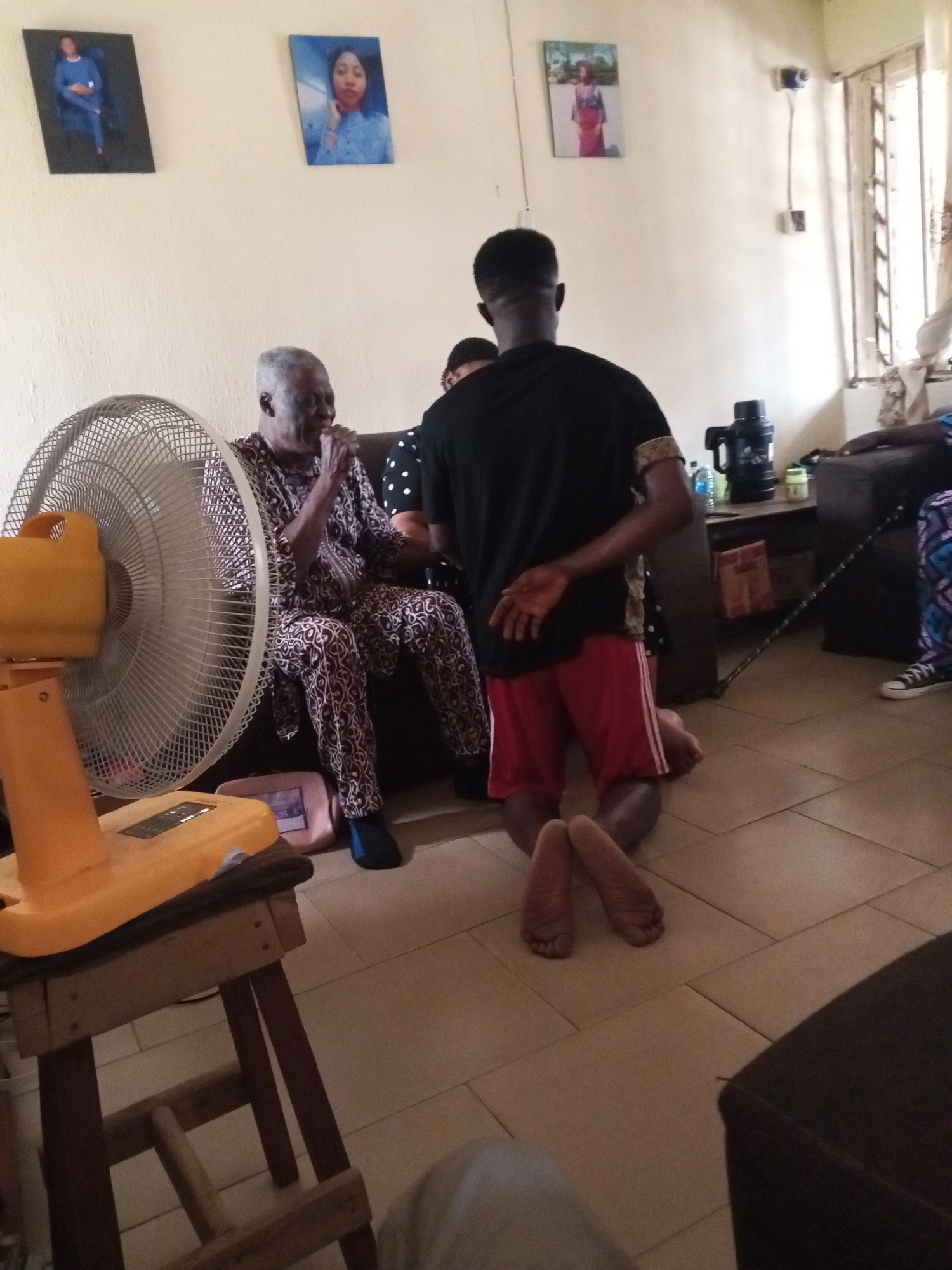 An indoor scene depicts a living room where two individuals are present. An elderly man, seated on a couch and wearing a patterned outfit, appears to be speaking with his hands clasped. Facing him, a young man is kneeling on the tiled floor with his back to the camera, wearing red shorts and a black t-shirt. A fan is positioned to the left of the frame, and a wooden stool supports its base. In the background, framed pictures hang on the wall, and a table with a kettle and other items is visible. The overall lighting suggests daylight filtering in from a window. The setting appears to be a residential interior.