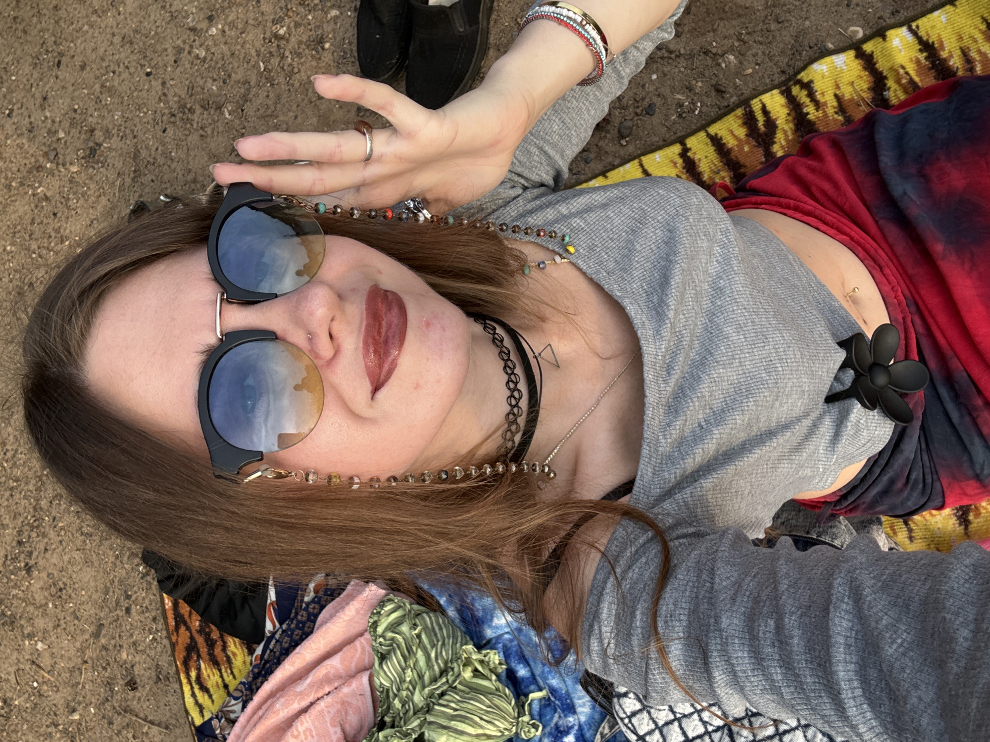 A woman lies on the ground, propped up on her elbows, with her head tilted towards the camera. She wears large, round sunglasses with a gradient lens, a gray long-sleeved crop top, and a red and black tie-dye skirt. A beaded chain is attached to her sunglasses, adorned with colorful beads and charms. She also wears a black choker necklace with a small silver triangle pendant, and several bracelets on her right wrist. Her left hand is raised to adjust her sunglasses. The background consists of the ground, with colorful fabrics and a pair of black shoes scattered around her.