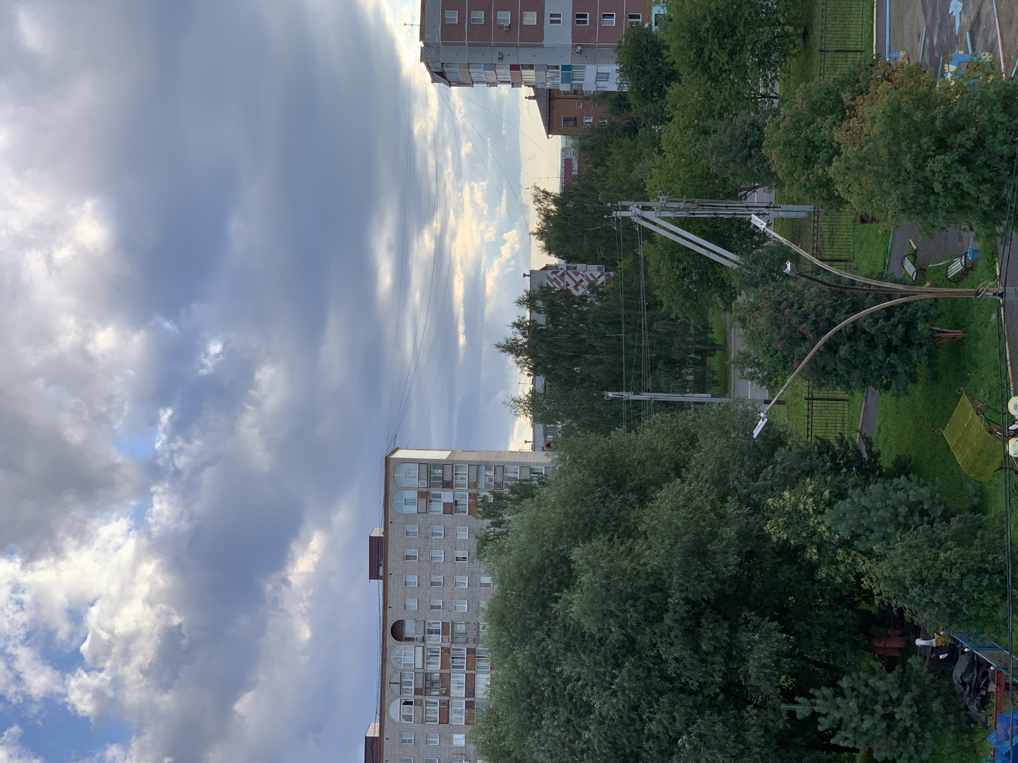 This is an exterior, eye-level, medium shot of a residential area in Prokopyevsk, Russia. The image is dominated by a cloudy sky with patches of sunlight breaking through. In the mid-ground, there are several multi-story apartment buildings, some with patterned facades and arched windows. Lush green trees and smaller evergreen trees are abundant throughout the scene, forming a dense canopy in places. A park or common area is visible with benches and a lamppost. The ground is covered in grass and paved areas. There are no people visible in the image. The overall impression is one of a typical urban landscape with significant greenery.