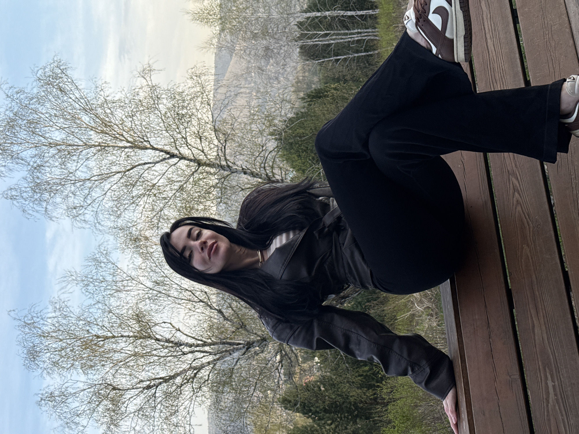 A woman is sitting cross-legged on a wooden structure. She is wearing a dark jacket, black pants, and brown and white sneakers. Her long black hair falls around her face. In the background, there are trees with sparse foliage, a mountain in the distance, and a sky with light clouds. The image was taken in the city of Krasnoyarsk, Russia.