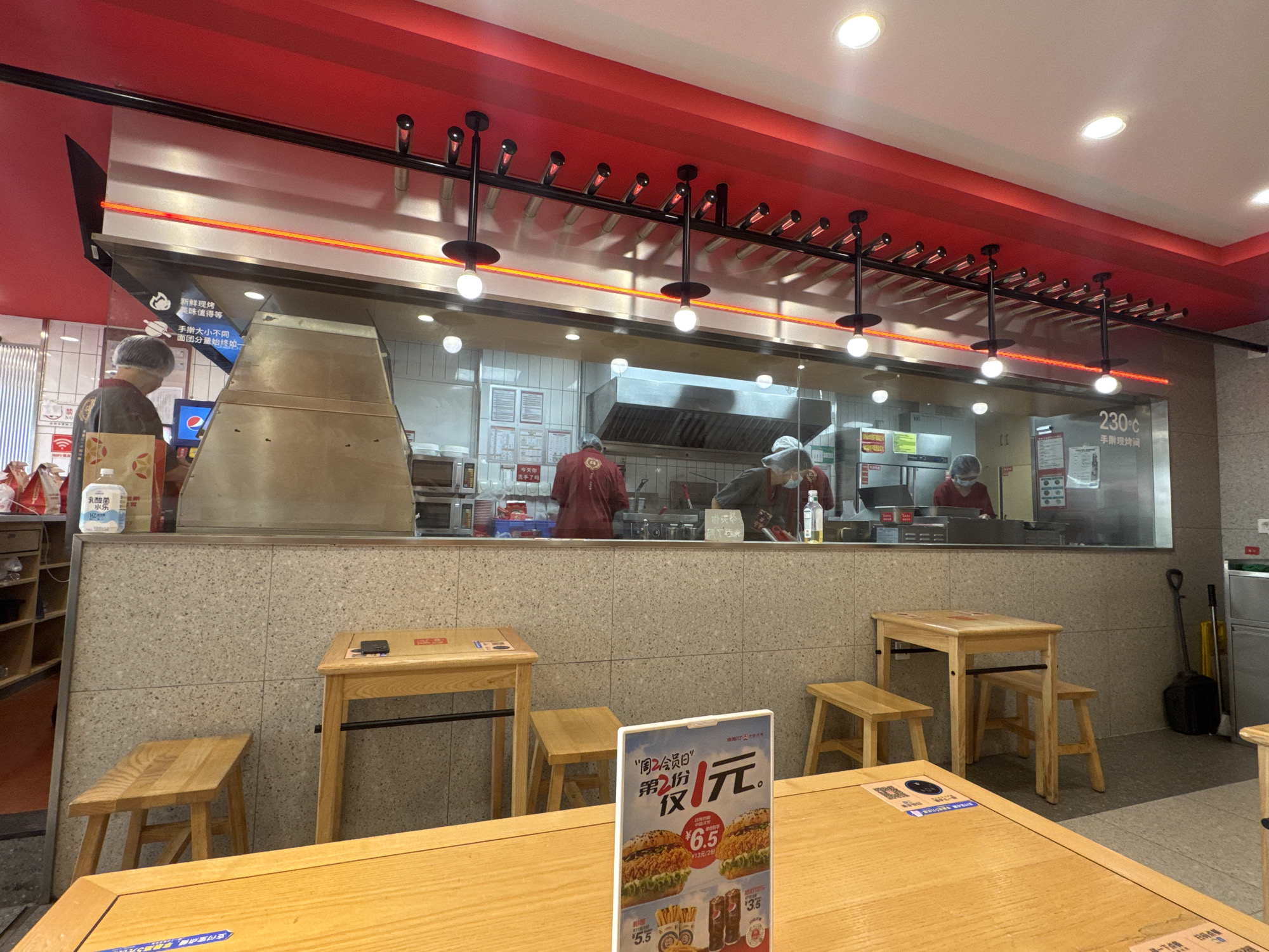 The image shows the interior of a fast-food restaurant, possibly a branch of a fried chicken chain, located in Dadong District, China. In the background, through a glass partition, several employees in red uniforms and hairnets are visible working behind a counter, preparing food in a kitchen environment. The kitchen area features stainless steel equipment, including a large range hood.
In the foreground, there are several wooden tables and stools arranged for customers. One table in the center has a menu displayed on it, showing pictures of burgers and fries, along with prices. Another table to the right has a small black object on it, possibly a remote control or a phone. The walls of the dining area are tiled with light-colored speckled tiles. The ceiling is painted red with overhead lighting fixtures. The overall atmosphere suggests a casual dining establishment.