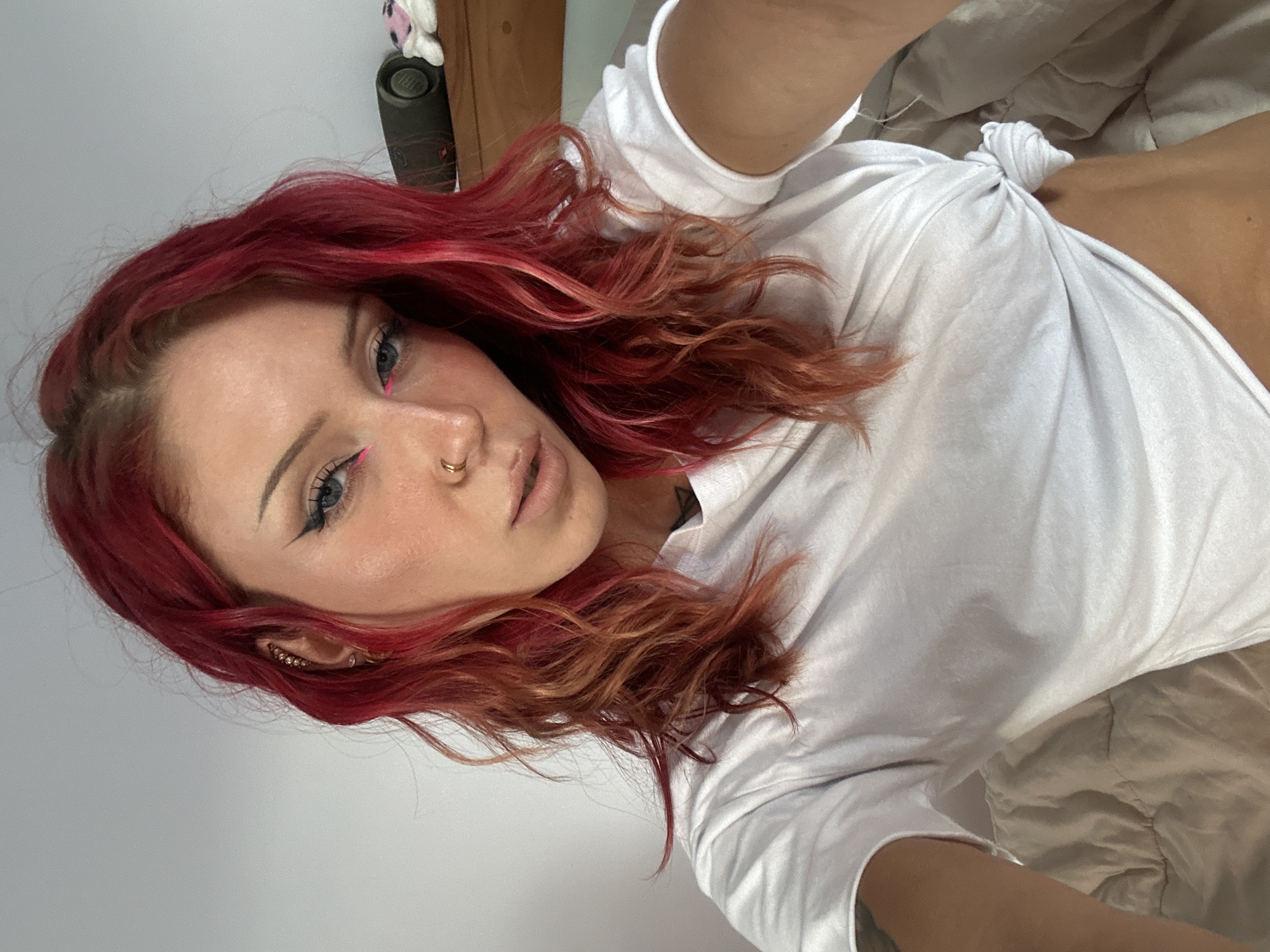 A woman with red hair is lying on a bed. She is wearing a white t-shirt that is tied in a knot. She has pink eyeliner on her upper eyelids and a nose ring. The location is a bedroom, the location in the city of North Kuta, Indonesia cannot be determined.