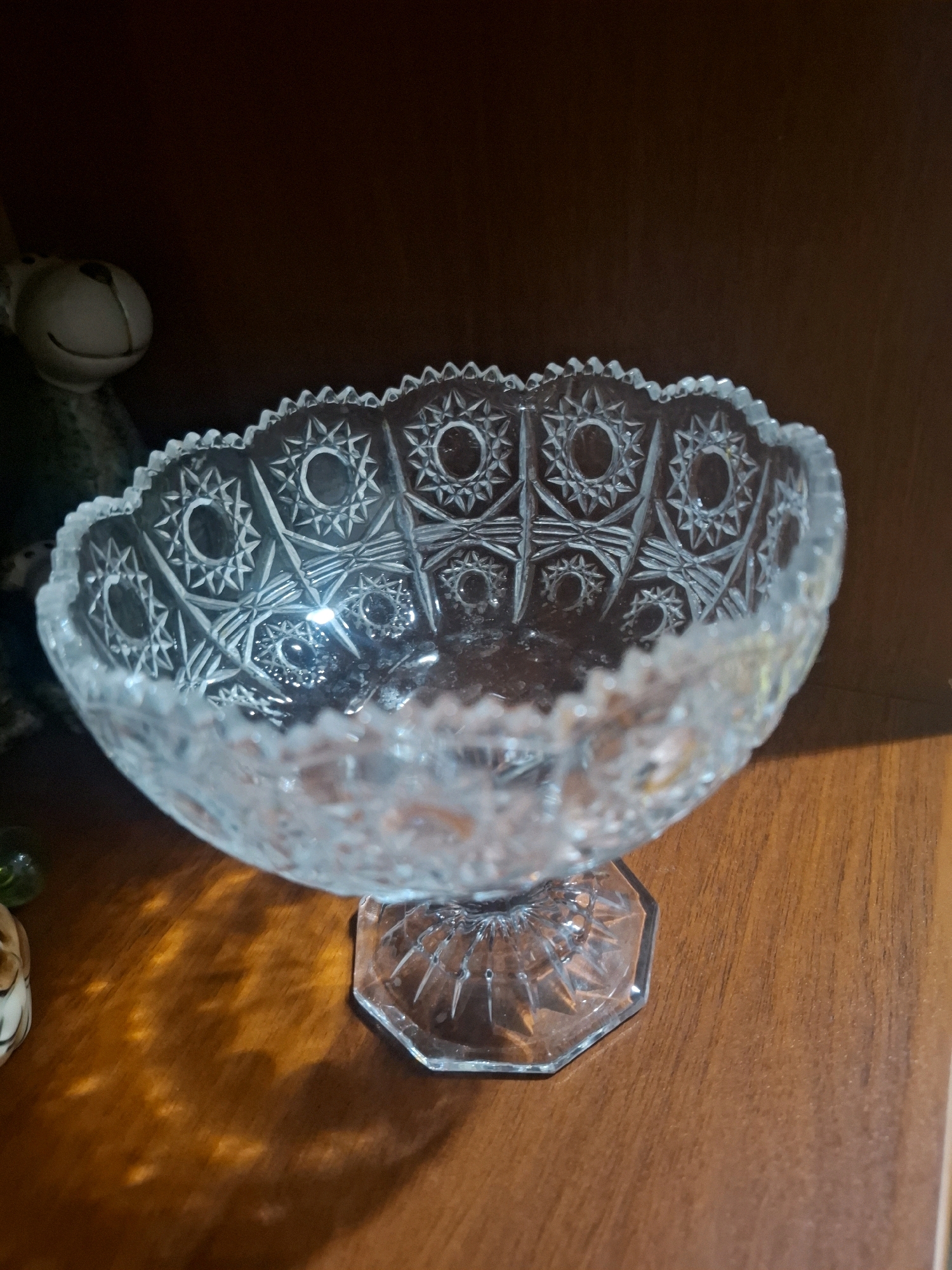 A clear, cut glass bowl with a decorative sawtooth rim sits on an octagonal glass pedestal. The bowl is intricately patterned with starburst designs. It is placed on a polished wooden surface, likely a table or shelf, which exhibits warm tones and wood grain. To the left of the bowl, partially visible, is the head of a light-colored, plush toy with dark eyes and a smiling mouth. In the background, a dark, indistinct surface suggests it is on a shelf in a room. The lighting creates dappled shadows on the wooden surface beneath the bowl. The scene is static and devoid of human presence. The location is not specified in the image.