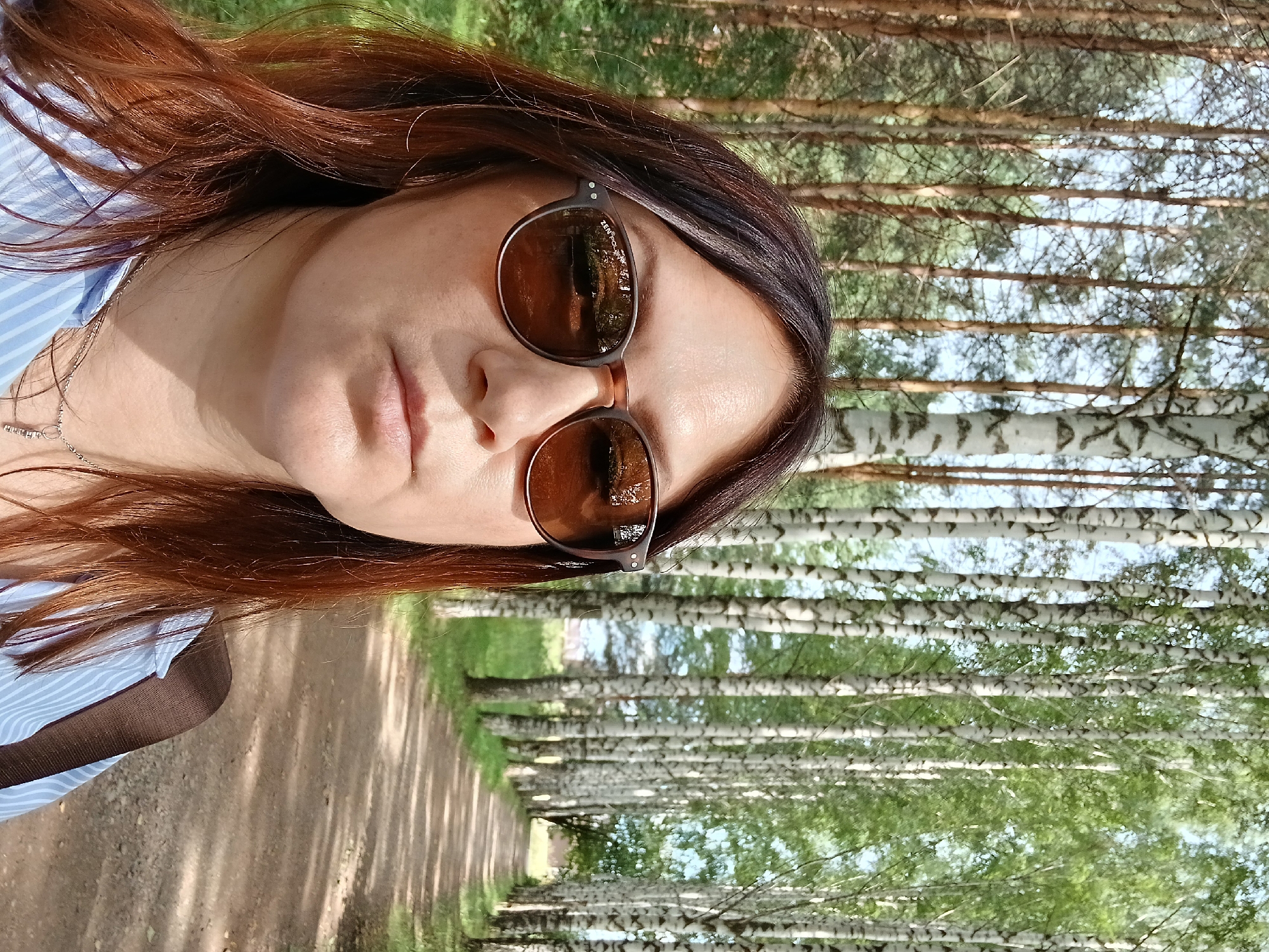 A woman with auburn hair, wearing sunglasses and a blue and white striped shirt, stands in a sun-dappled forest path lined with birch trees. She is looking into the camera, and the reflection of the trees is visible in her sunglasses. The path appears to be made of dirt and is surrounded by greenery. The location is the birch alley in the city of Osa, Russia.