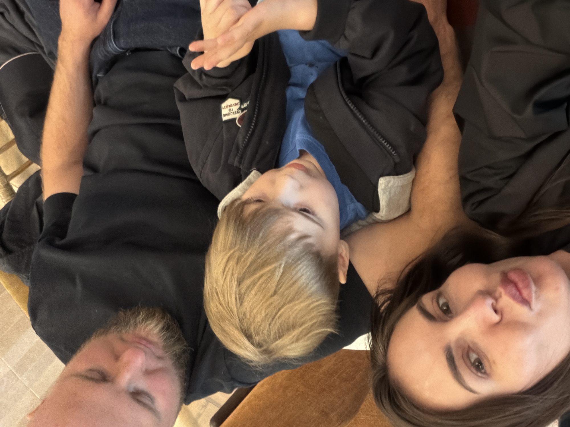 A close-up, angled selfie shows three people lying down and looking at the camera. The person on the left is a man with fair skin and a beard, wearing a black t-shirt. In the center is a young child with blond hair, wearing a blue shirt and a black jacket. The person on the right is a woman with fair skin and dark brown hair, wearing a black top. The environment is indistinct, but a wooden floor or furniture is visible in the background. The location is unknown and there is nothing to suggest that the picture was taken in the city of Krasnoyarsk, Russia.