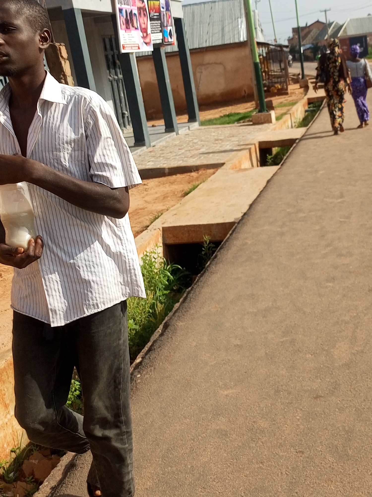 The image depicts a man standing on a paved path, holding a transparent plastic bottle filled with a white substance. He is wearing a white collared shirt with thin vertical dark stripes and dark pants. To his left, a building with a grey and white facade is visible, featuring posters on its exterior. Further in the background, two women are walking away from the camera on a different path, one wearing a colorful patterned dress and the other in a purple headscarf and matching skirt. The environment suggests an outdoor urban or semi-urban setting with some greenery and concrete structures.