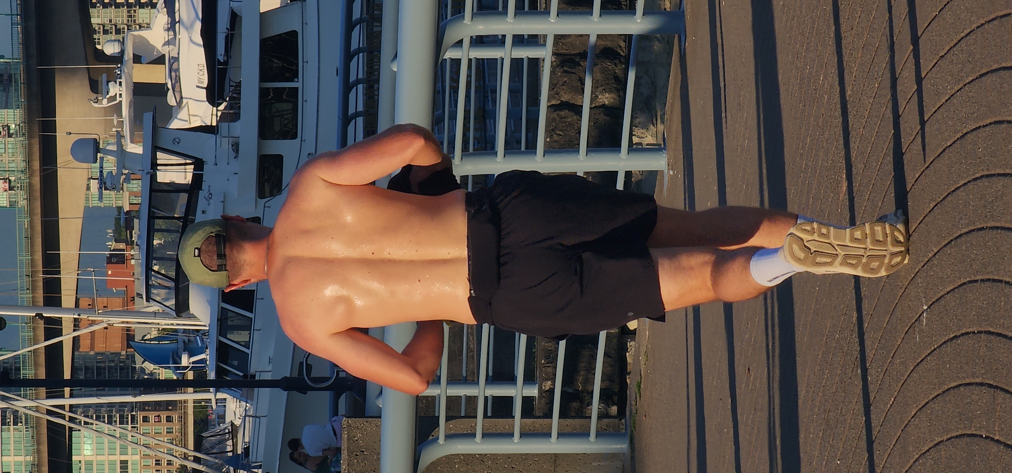 A bare-chested man wearing black shorts, a green baseball cap, white socks, and beige athletic shoes is walking away from the camera on a paved walkway with a repeating curved pattern. He has a dark belt or pouch around his waist. His back is glistening with sweat. In the background, there is a metal railing with blue uprights and horizontal bars, and beyond that, several white yachts are docked in a harbor. Further in the background, there are city buildings and a bridge with visible traffic. Two people are seated on a concrete structure to the left of the frame, partially visible.