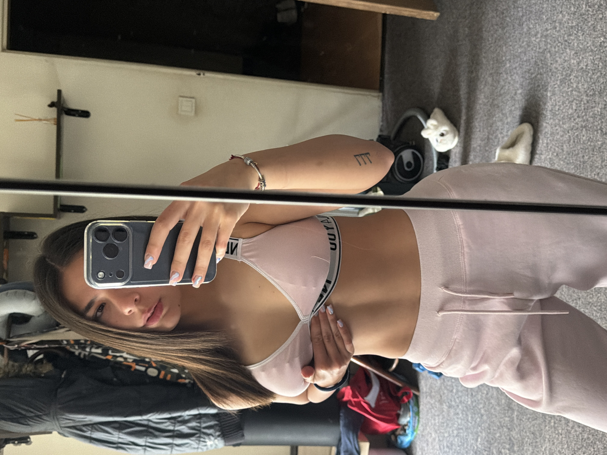 A young woman is taking a selfie in a mirror. She is wearing a pink sports bra and pink sweatpants. The background appears to be a room with various objects, including clothes and a vacuum cleaner. There is no indication of the location being in Gabrovo, Bulgaria.